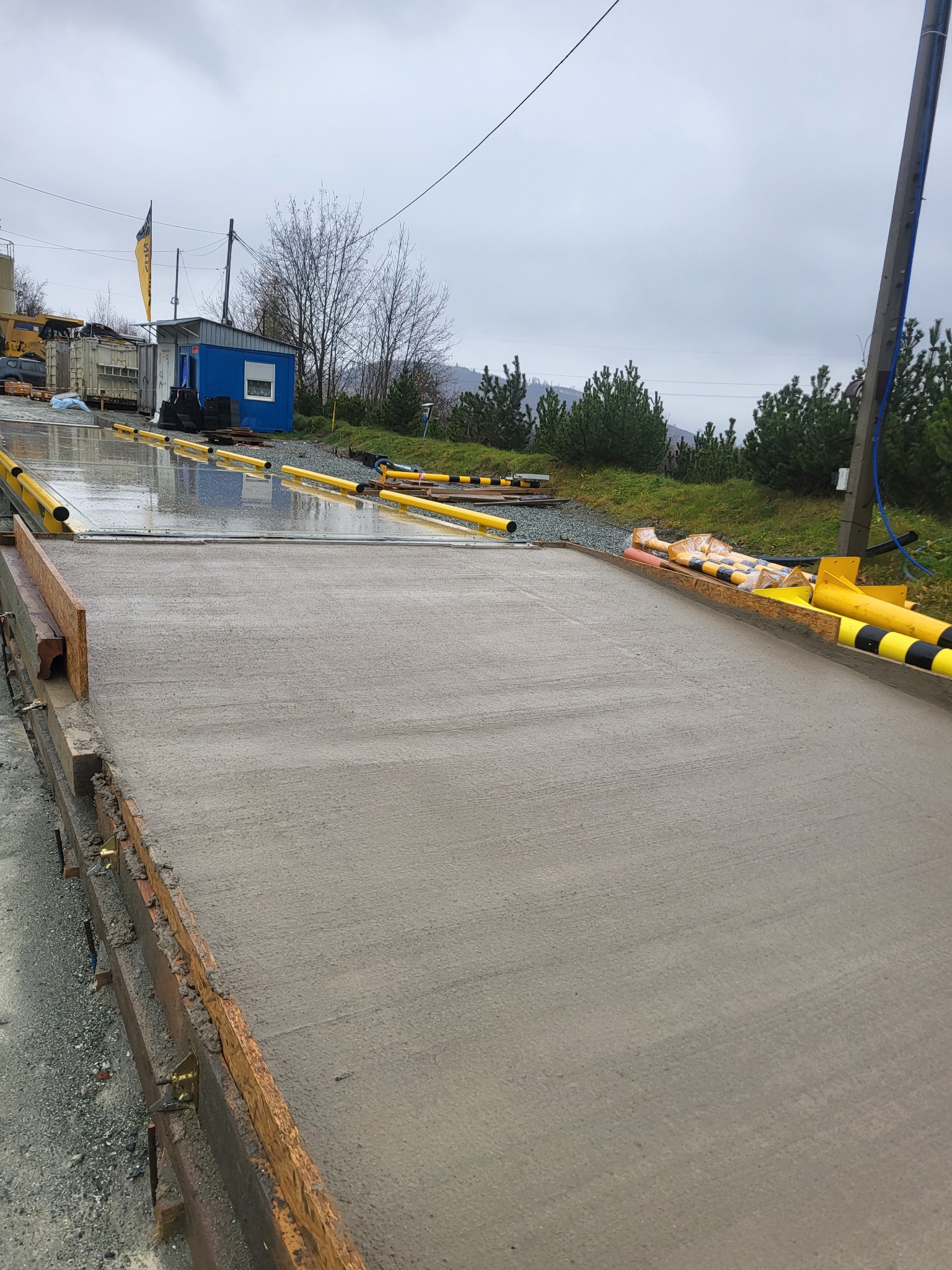 Świeżo wylany, mokry beton w szalunku, widok z góry. W tle budka, drzewa i pochmurne niebo. Żółte elementy zabezpieczające na krawędziach.