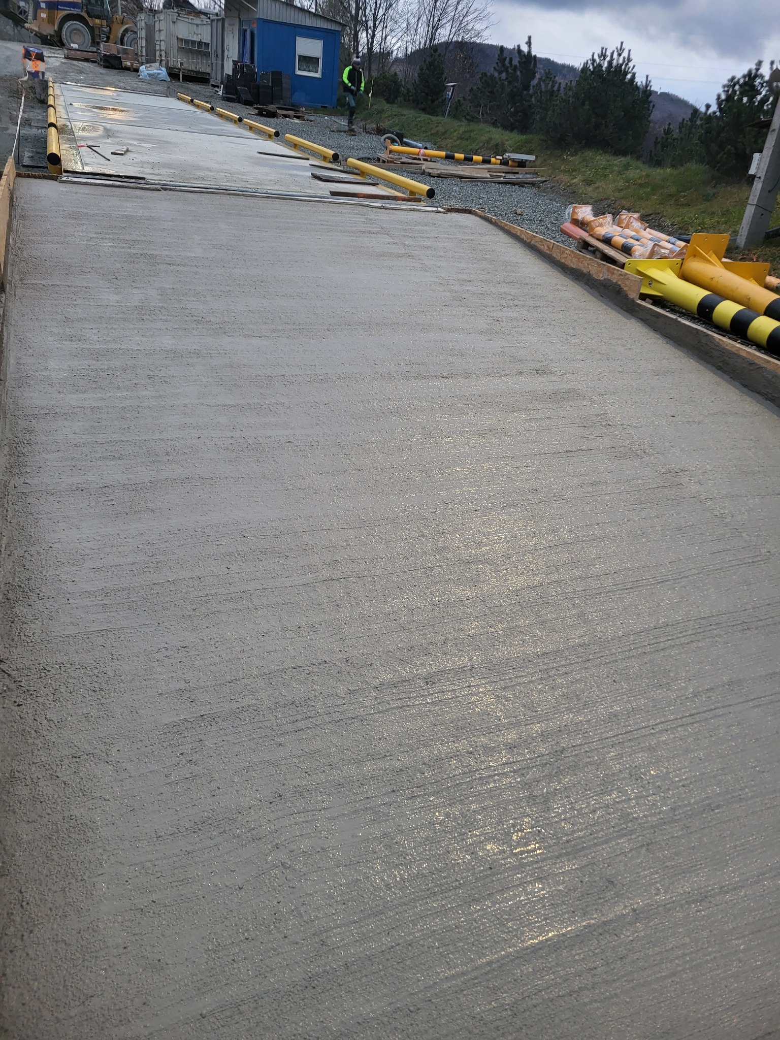 Świeżo wylany, gładki betonowy fundament z żółtymi rurami ochronnymi na obrzeżach, w tle budynek i zieleń. Widok z góry, pochmurne niebo.