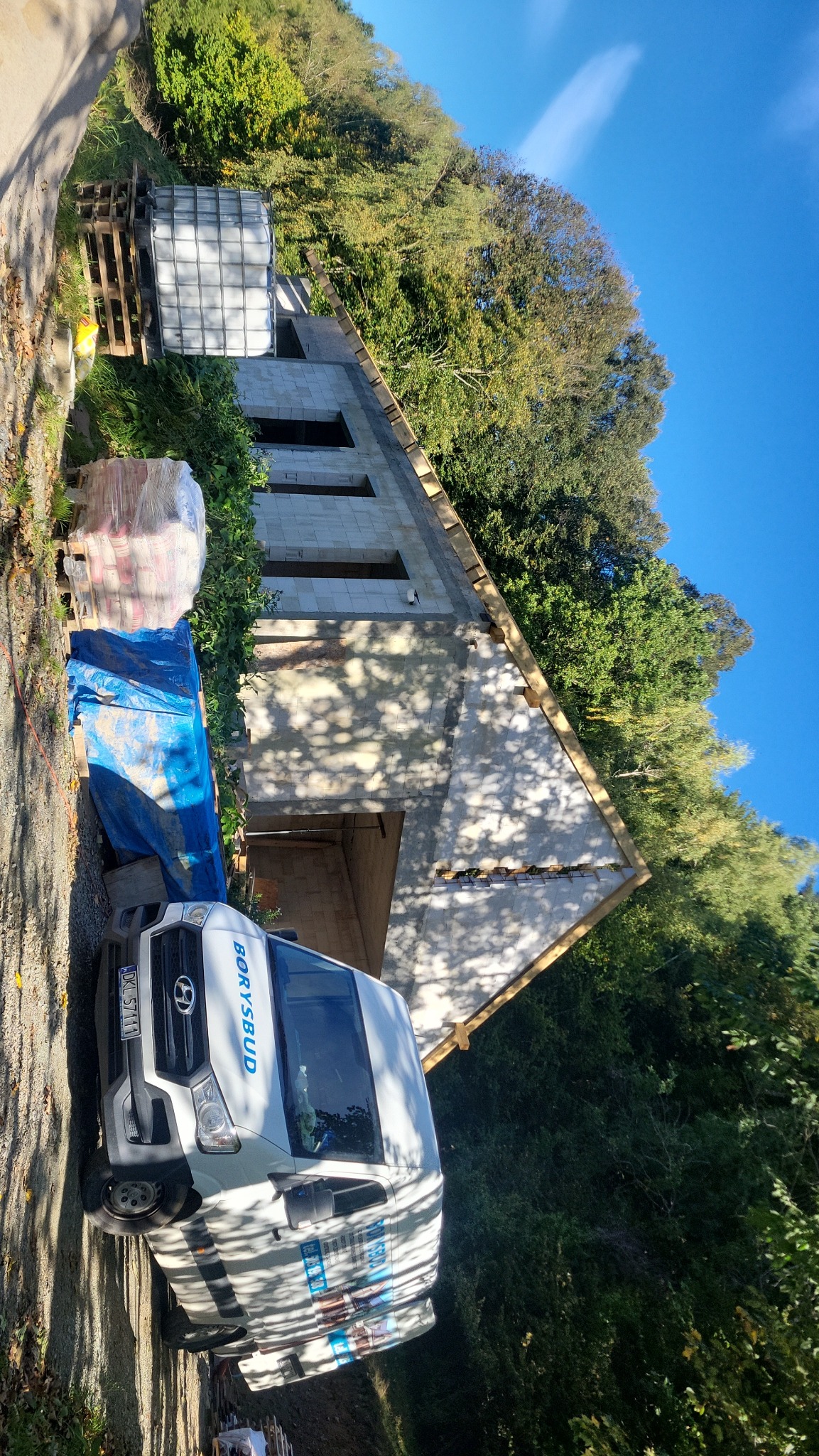 Budowa domu z keramzytu w trakcie: ściany wzniesione, widoczny szkielet dachu, palety z materiałami i firmowy bus Borysbud na tle zieleni i błękitnego nieba.