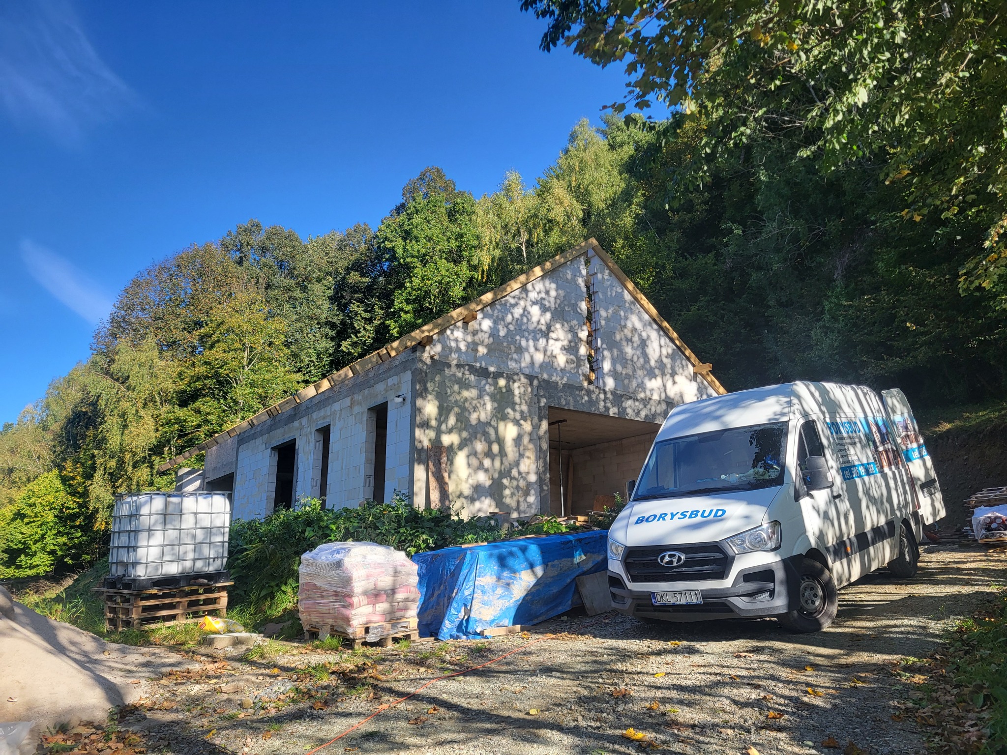 Budowa domu z keramzytu w trakcie: ściany wzniesione, więźba dachowa, materiały budowlane obok i firmowy bus Borysbud na tle zieleni. Słoneczny dzień.