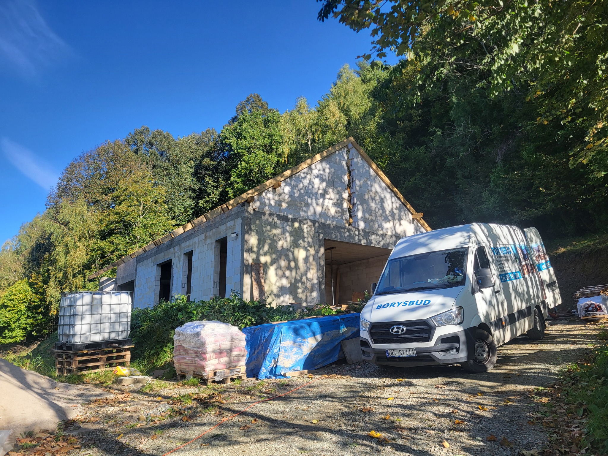 Budowa domu z pustaków: surowy stan, widoczny szkielet dachu, materiały budowlane na paletach, firmowy bus Borysbud na tle zieleni i błękitnego nieba.