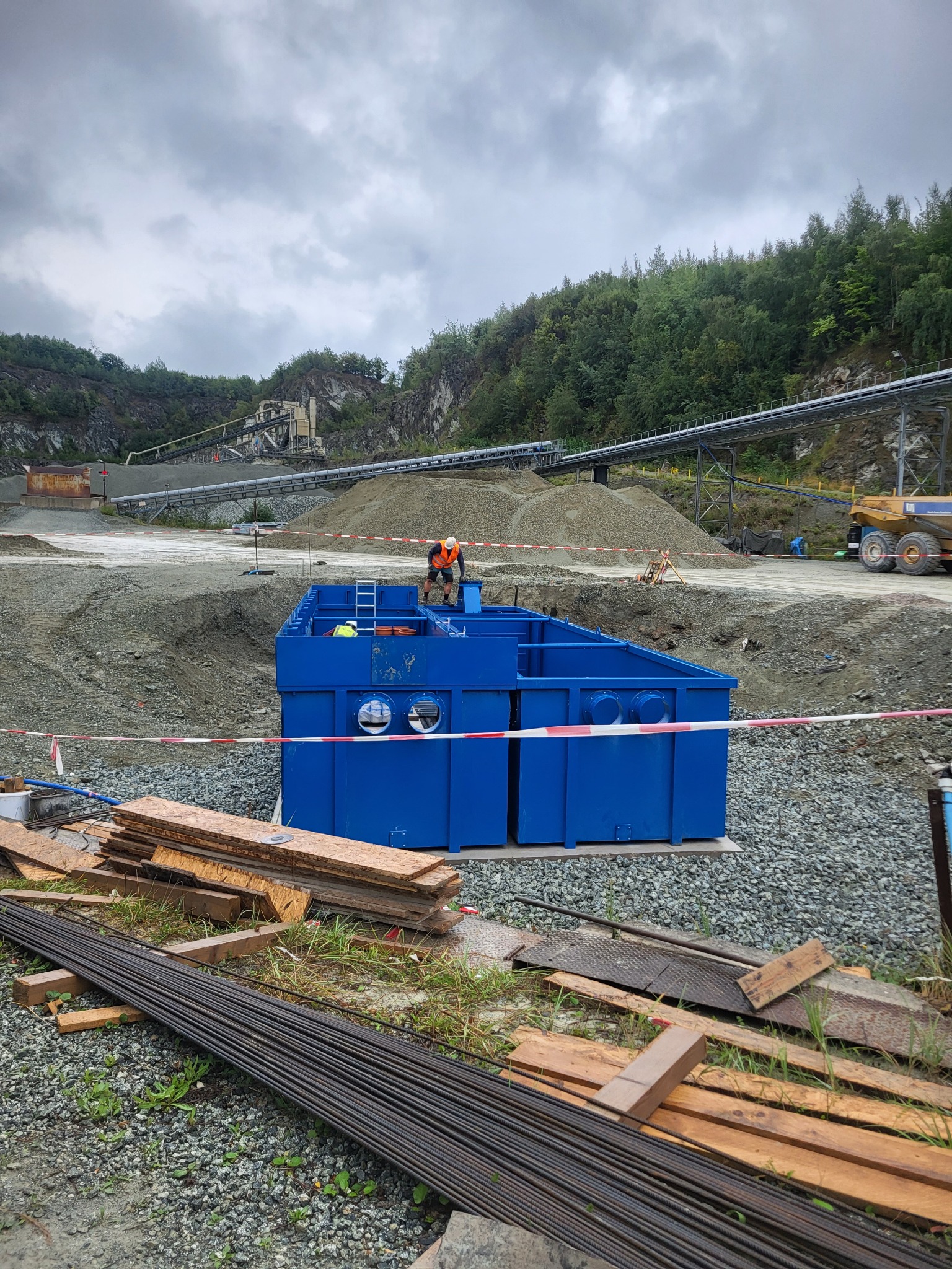 Niebieska konstrukcja w wykopie, pracownicy w kaskach, taśmy ostrzegawcze, teren budowy z transportem kruszywa w tle. Górniczy krajobraz.