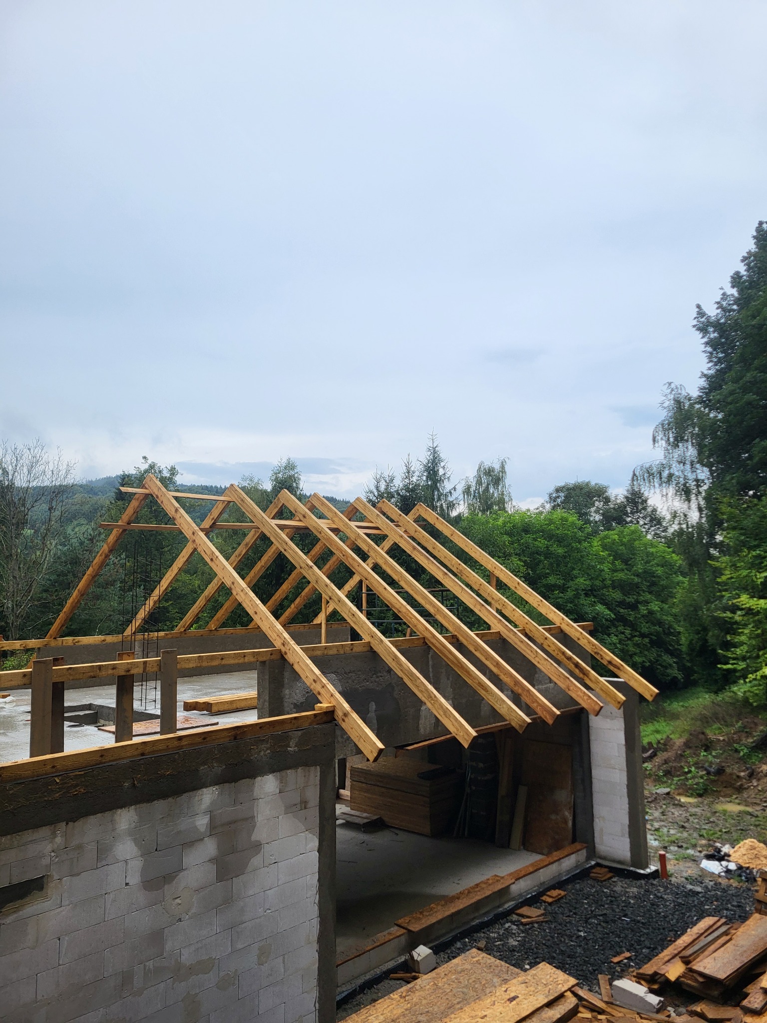 Wznoszenie konstrukcji dachu z drewnianych belek na surowym budynku z betonu komórkowego, w tle zalesione wzgórza pod pochmurnym niebem.