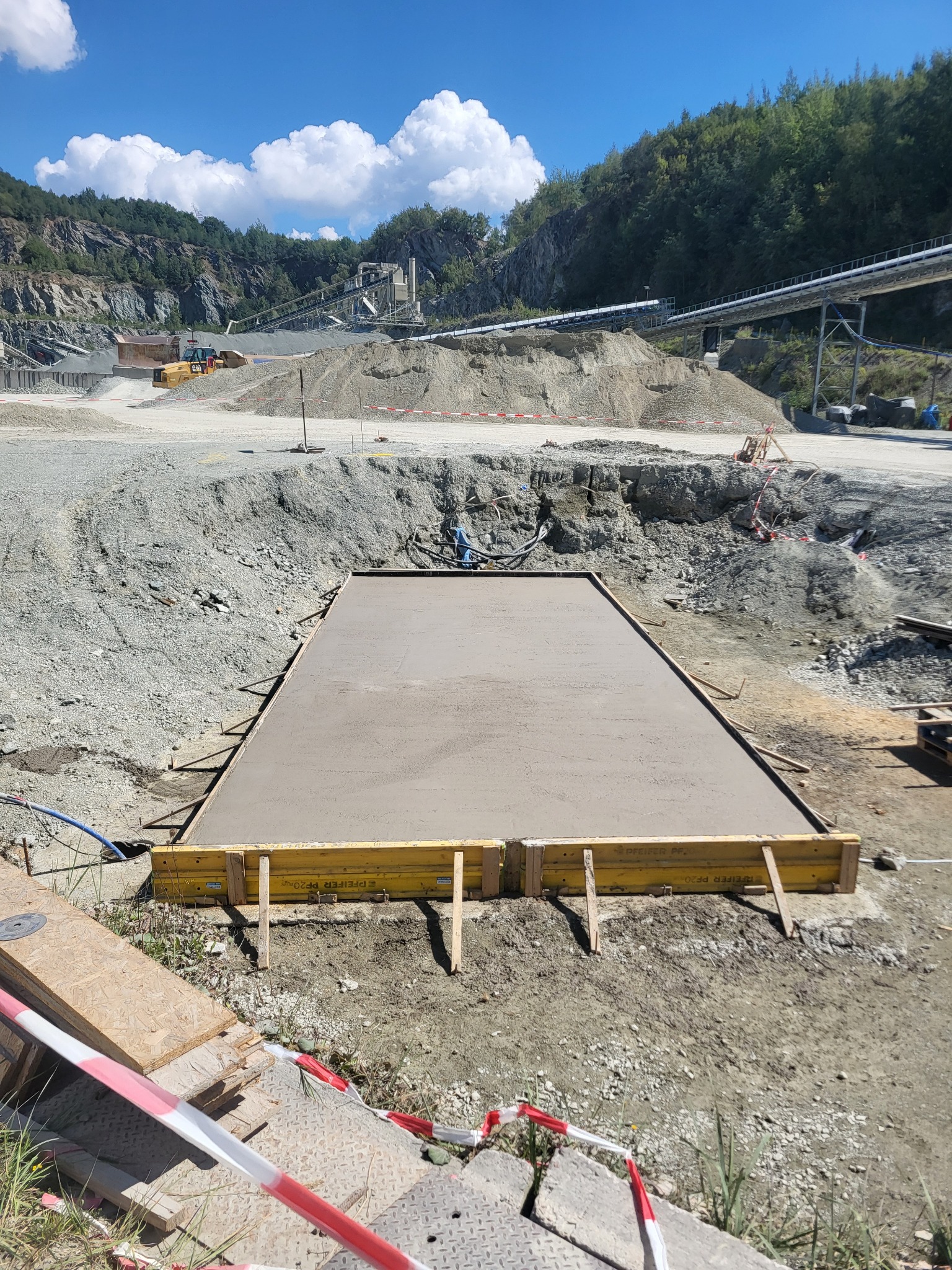 Wylany fundament w szalunku na tle kamieniołomu. Widoczne deski szalunkowe, świeży beton i otoczenie placu budowy z maszynami i hałdami ziemi.