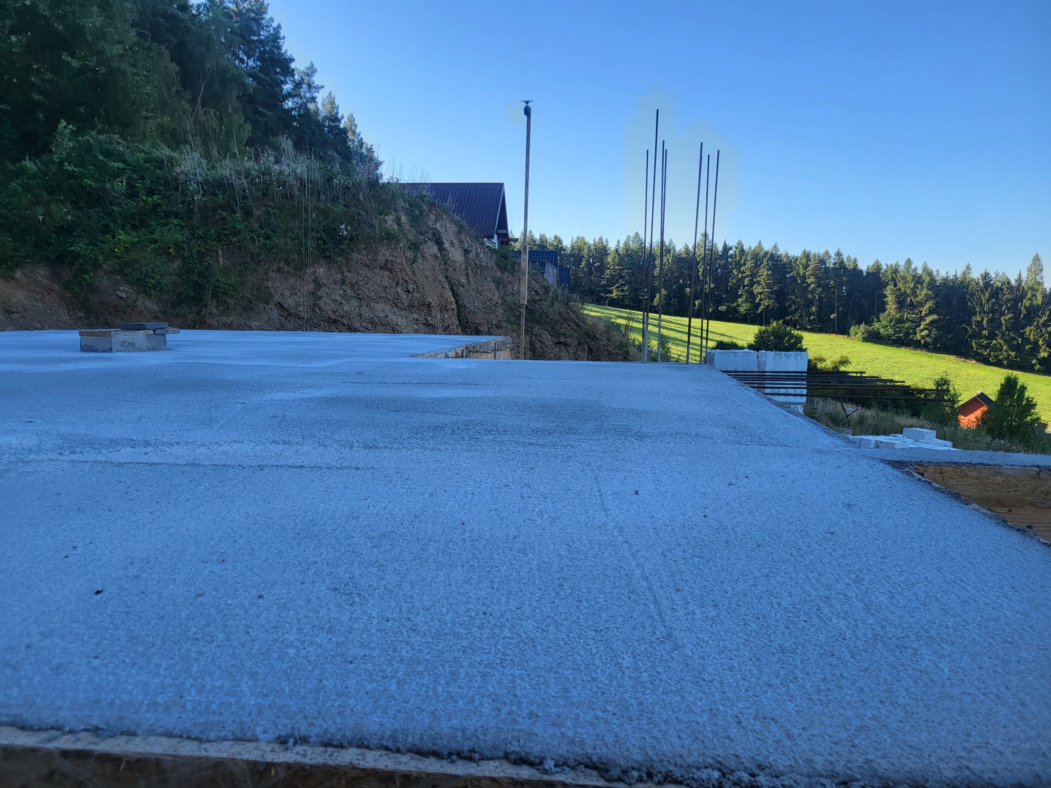 Wylana płyta fundamentowa z wystającymi prętami zbrojeniowymi na tle zieleni w Nowej Rudzie. Widok na wzgórze i las.