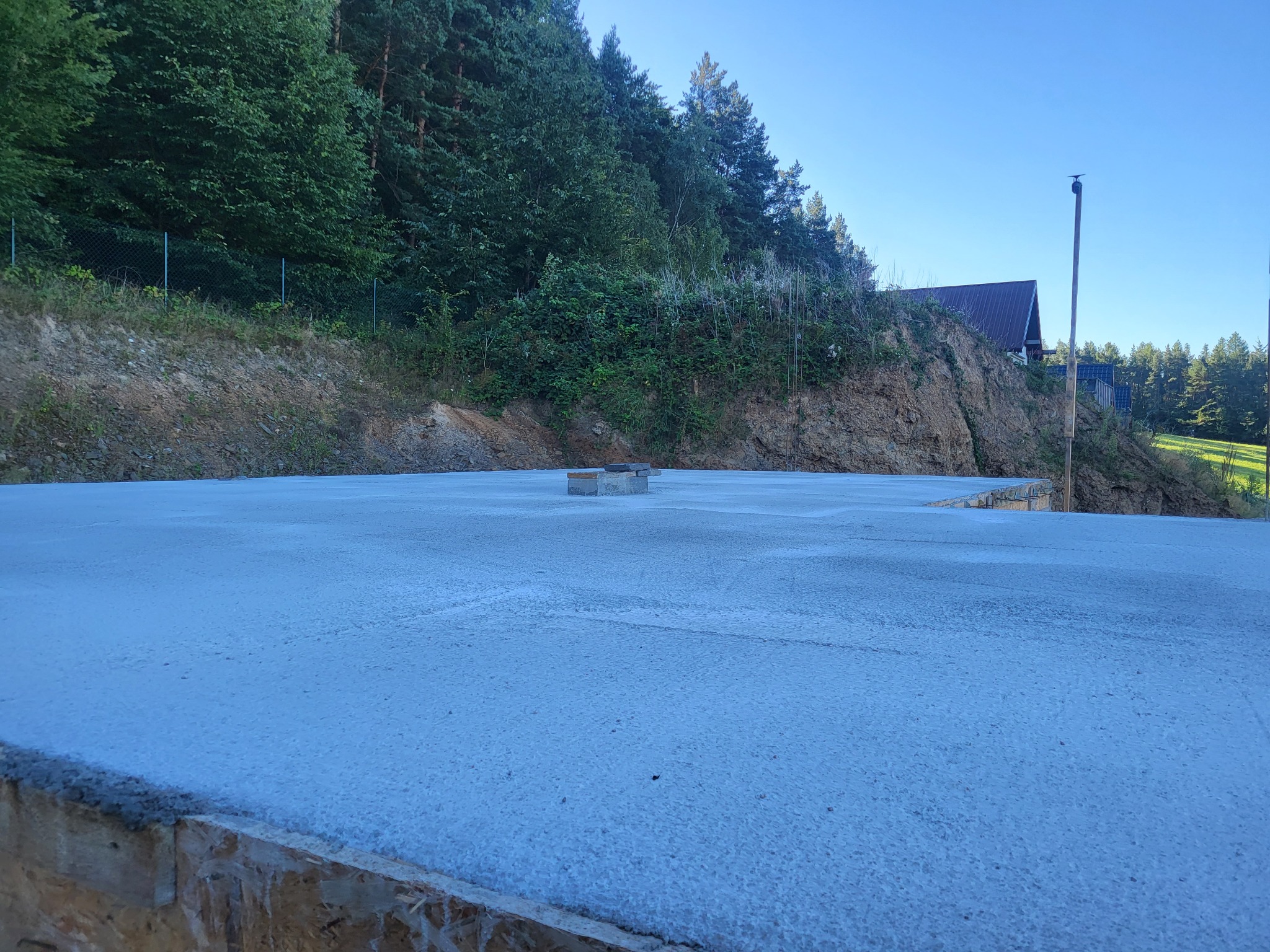 Wylana płyta fundamentowa na tle zieleni i błękitnego nieba. Widoczne szalunki i wystające elementy konstrukcyjne. Ujęcie z niskiej perspektywy.