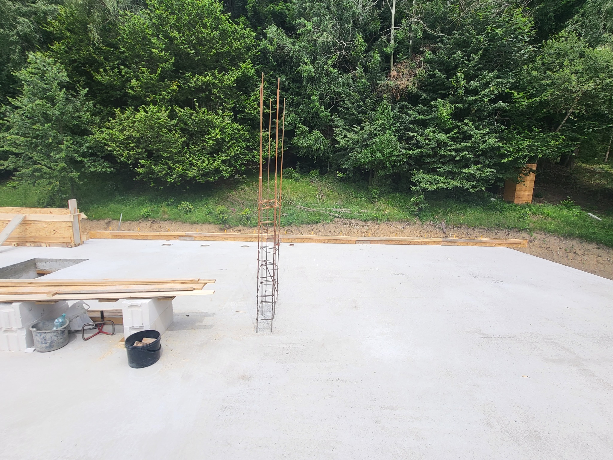 Zalana płyta fundamentowa z wystającą konstrukcją zbrojeniową na tle zieleni. Widoczne szalunki i materiały budowlane. Prace fundamentowe w toku.