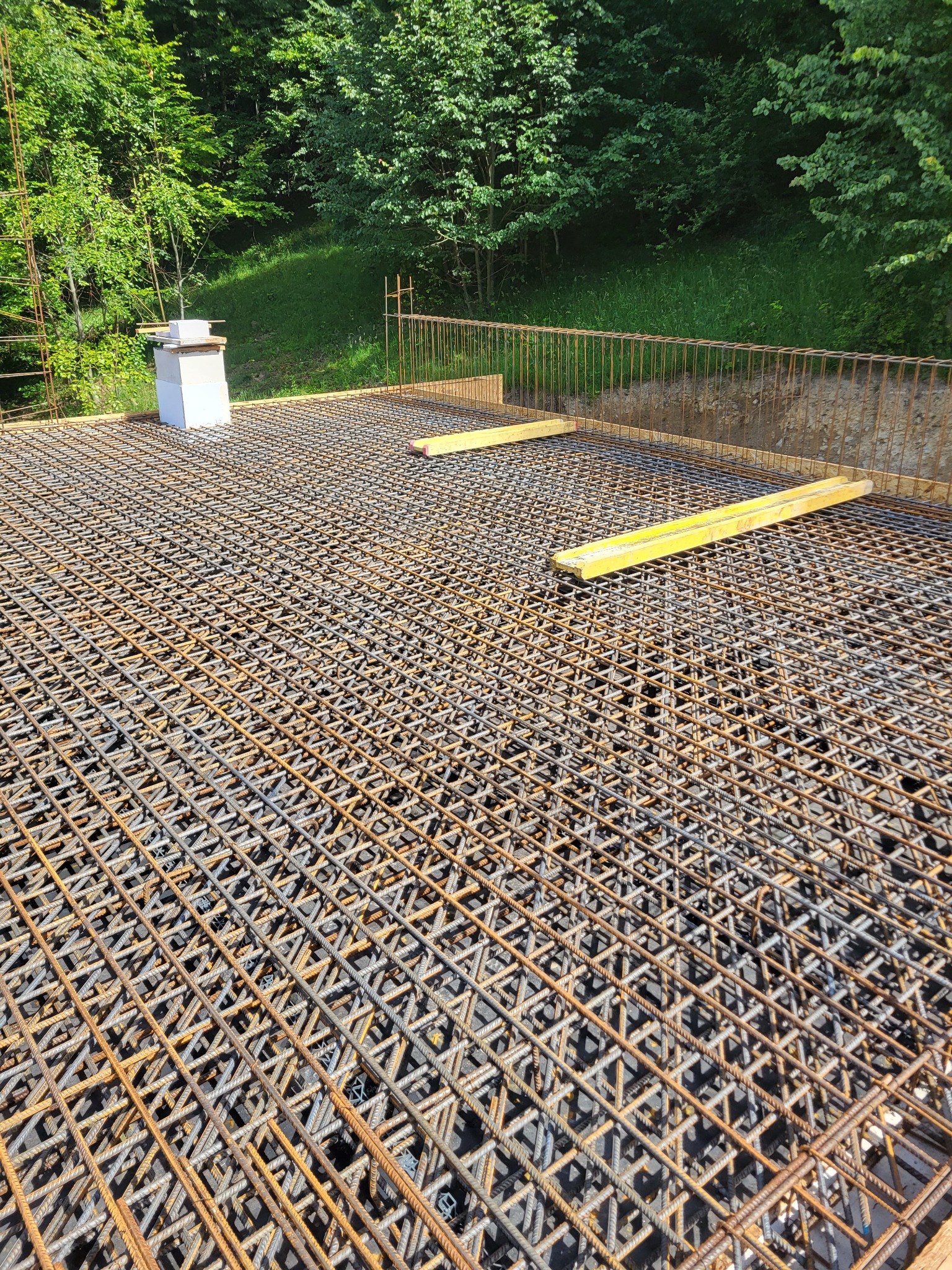 Zbrojenie płyty fundamentowej z widocznymi elementami szalunku i zielonym tłem drzew. Dwa żółte elementy na zbrojeniu. Widok z góry na siatkę zbrojeniową.