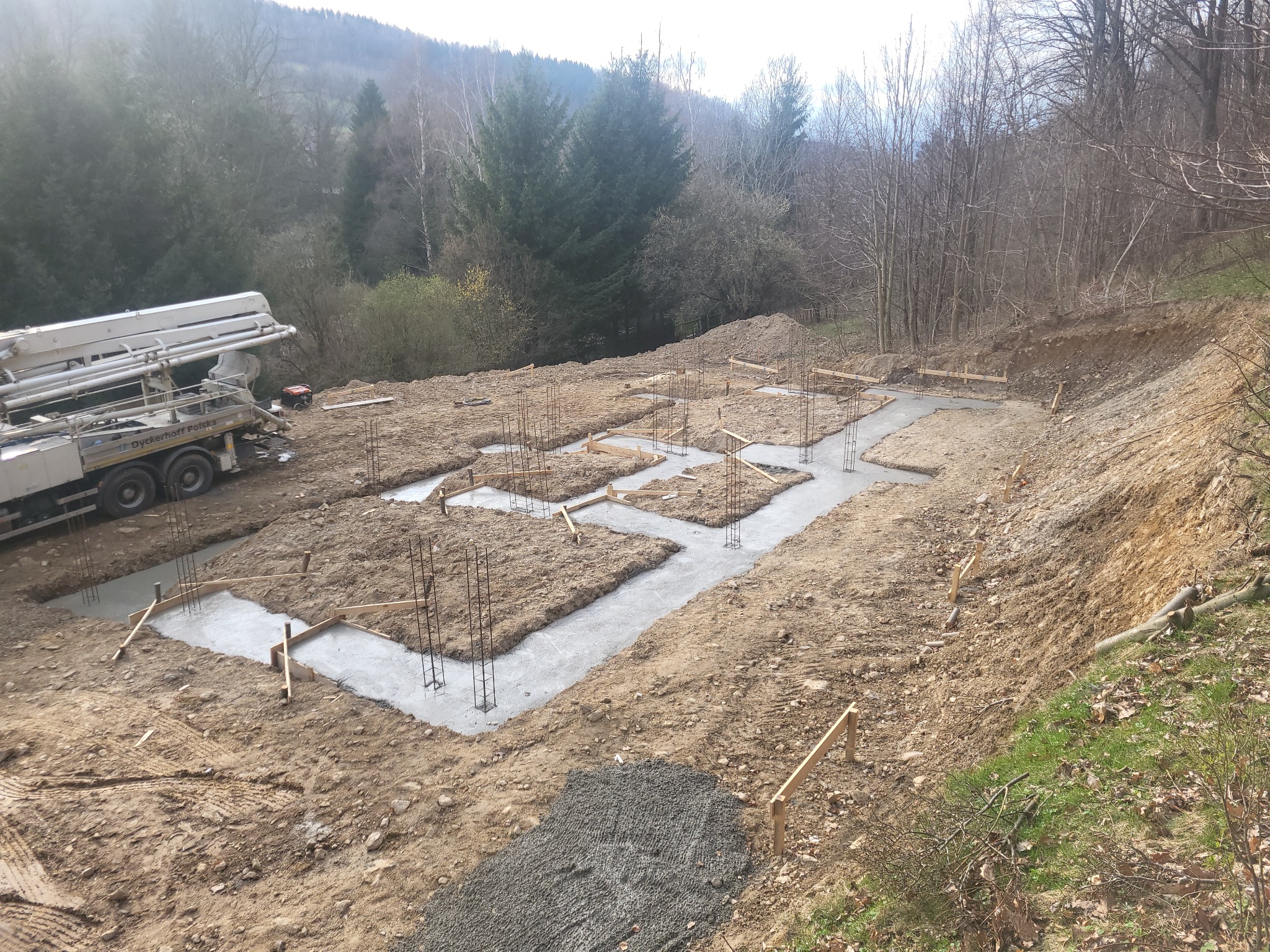 Wylane fundamenty pod budowę domu na nierównym terenie, widoczny betonowy szalunek i zbrojenie, w tle las i pompa do betonu.