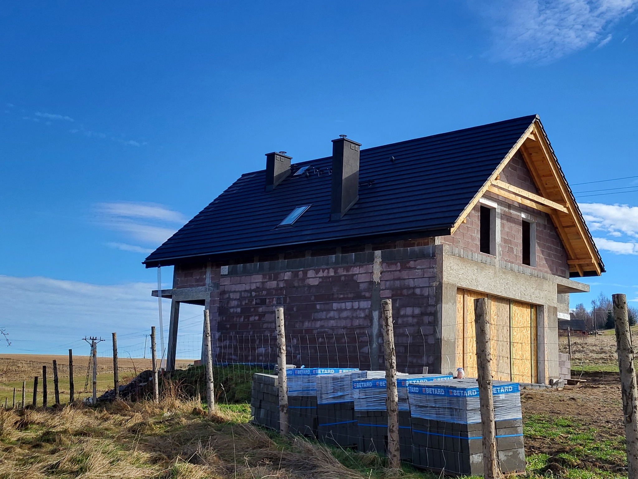 Dom w trakcie budowy z ciemną dachówką i oknem dachowym, widoczne palety z bloczkami, ogrodzenie z drutu i słupków, błękitne niebo.