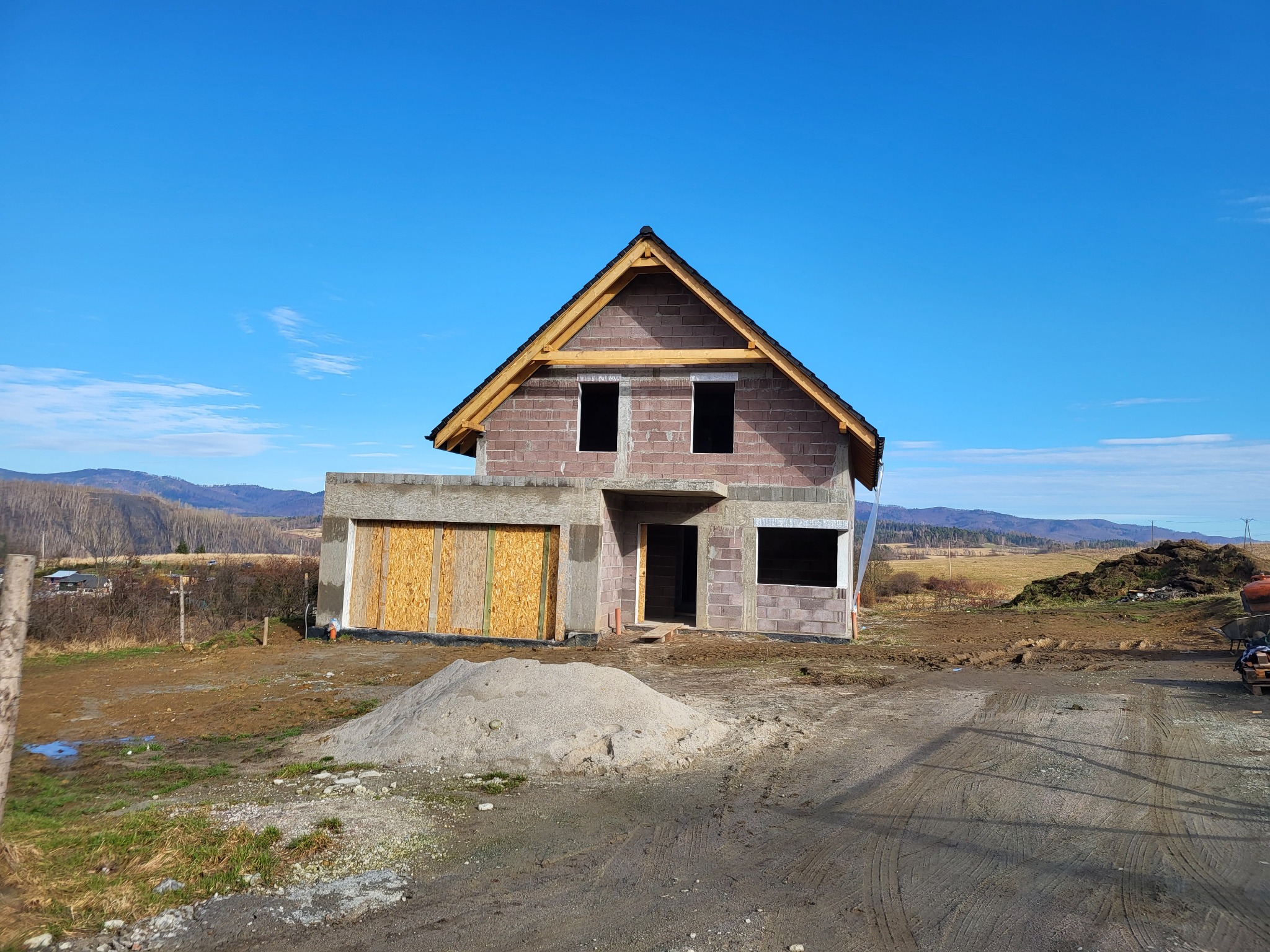 Niedokończony dom z keramzytu w stanie surowym otwartym, z widocznym drewnianym szkieletem dachu i oknami bez szyb, na tle pagórkowatego krajobrazu w słoneczny dzień.