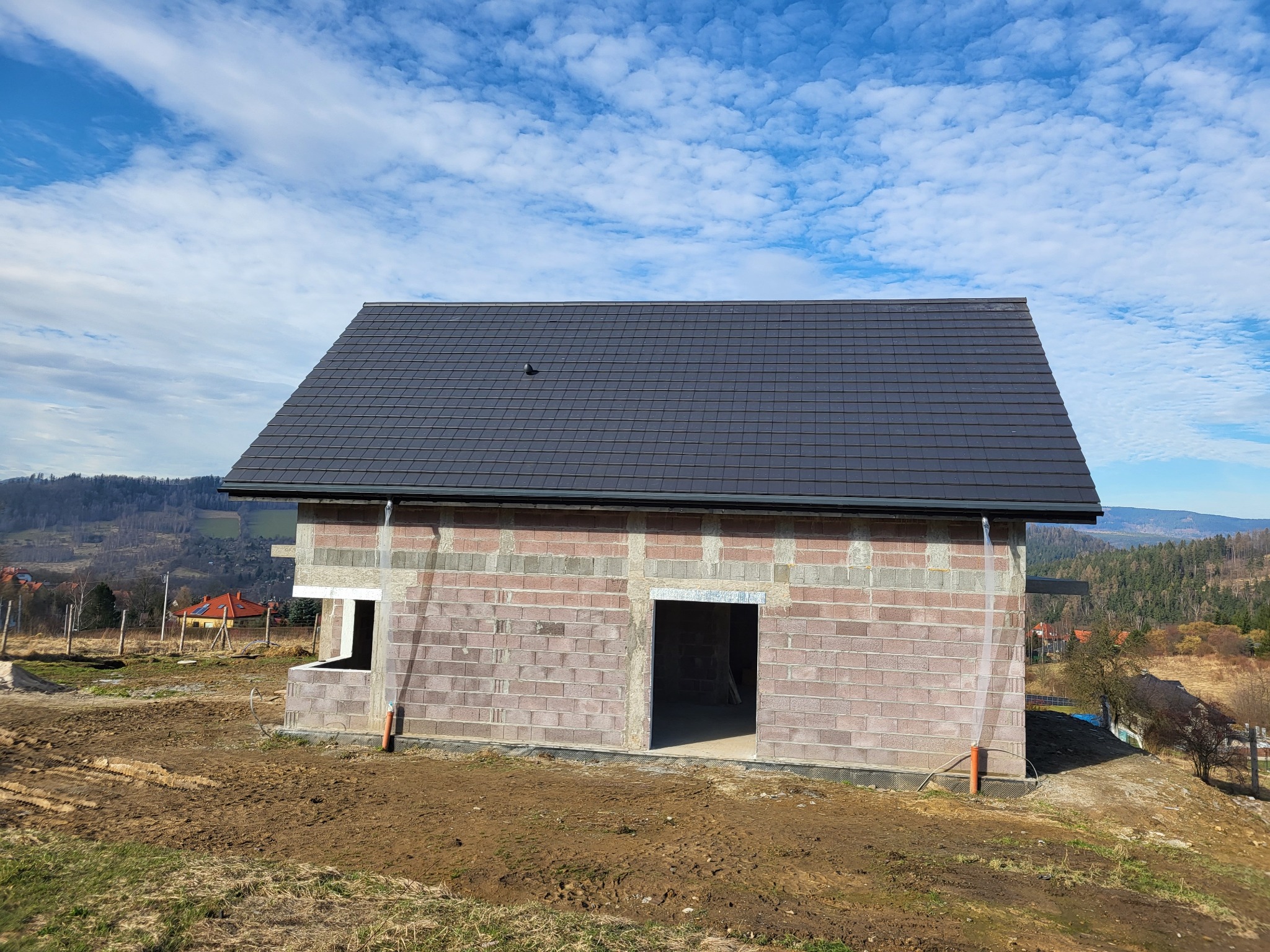 Niedokończony dom z keramzytu z ciemnym dachem na tle pagórkowatego krajobrazu i błękitnego nieba z chmurami. Widoczne surowe ściany i otwory okienne.