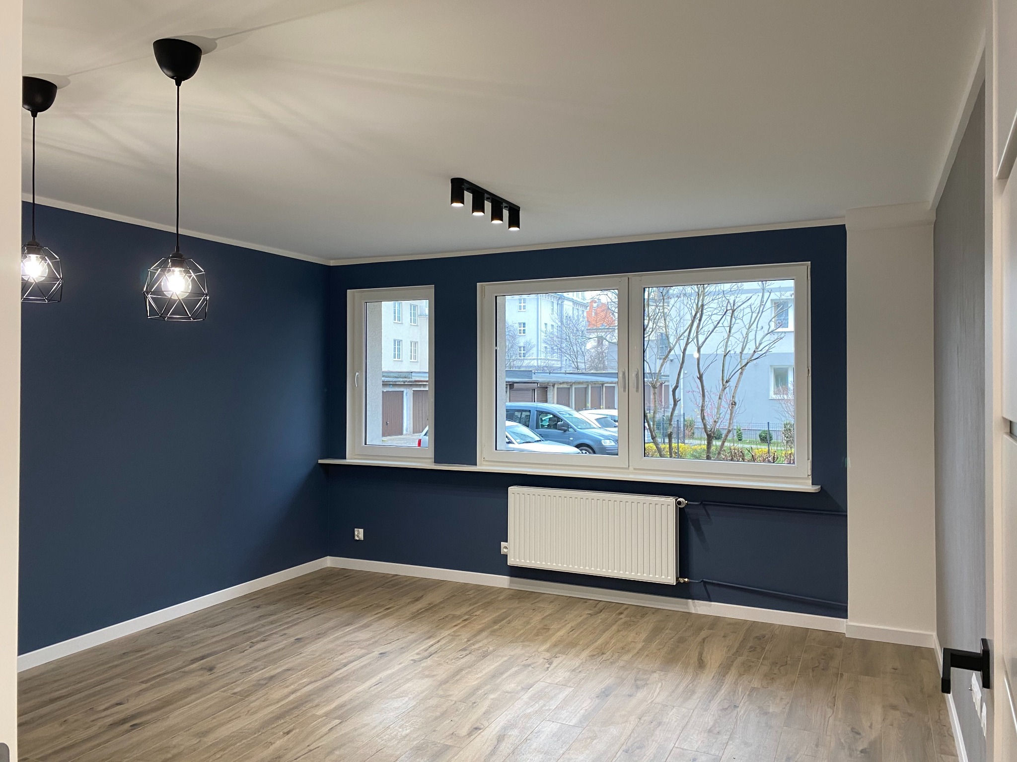 Wykończone wnętrze pokoju z drewnianą podłogą, granatową ścianą, białymi oknami i grzejnikiem. Widoczne lampy sufitowe i listwy przypodłogowe.