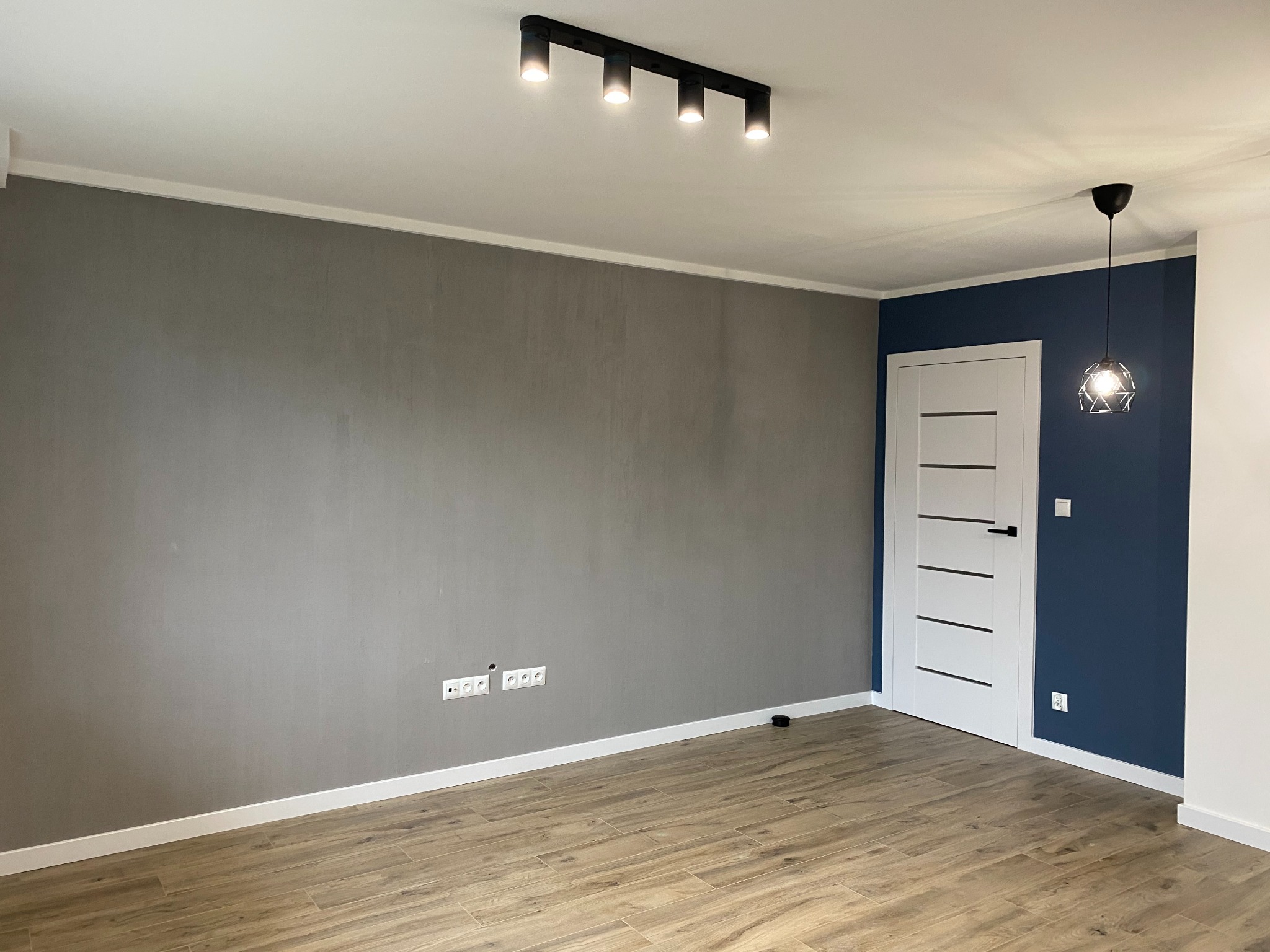 Wnętrze po remoncie: szara ściana, drewnopodobna podłoga, białe listwy przypodłogowe, niebieska ściana z białą stolarką drzwiową i nowoczesnym oświetleniem.