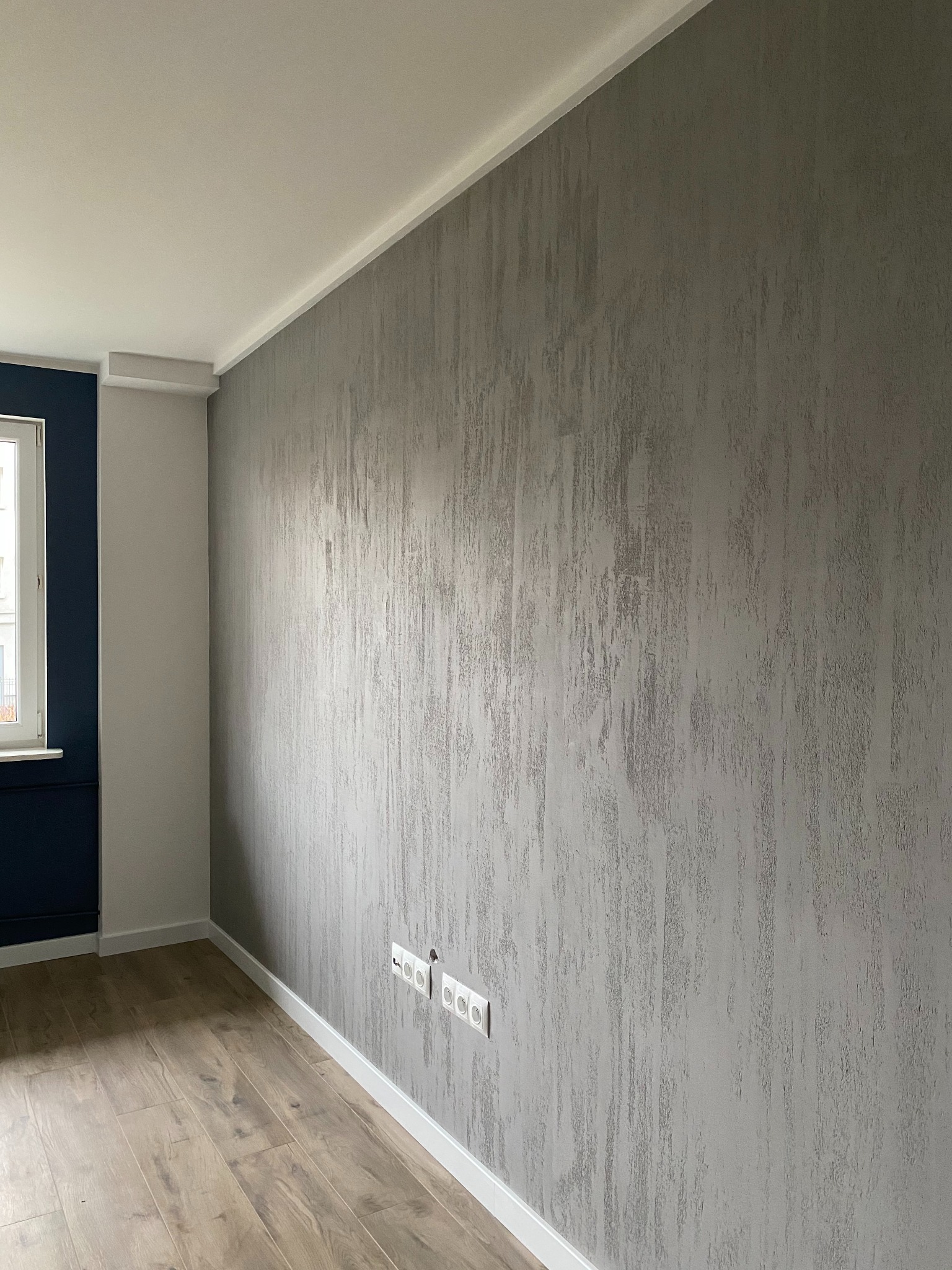 Pokój po remoncie: szara ściana z fakturą, drewniana podłoga, białe listwy przypodłogowe i okno w ciemnej ramie. Widoczne gniazdka elektryczne.