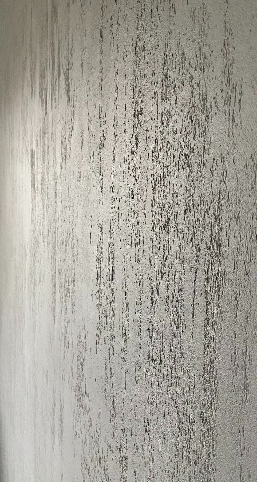 Szara ściana z dekoracyjną fakturą, imitującą pionowe smugi. Neutralna kolorystyka, delikatny wzór, widoczne detale struktury tynku.