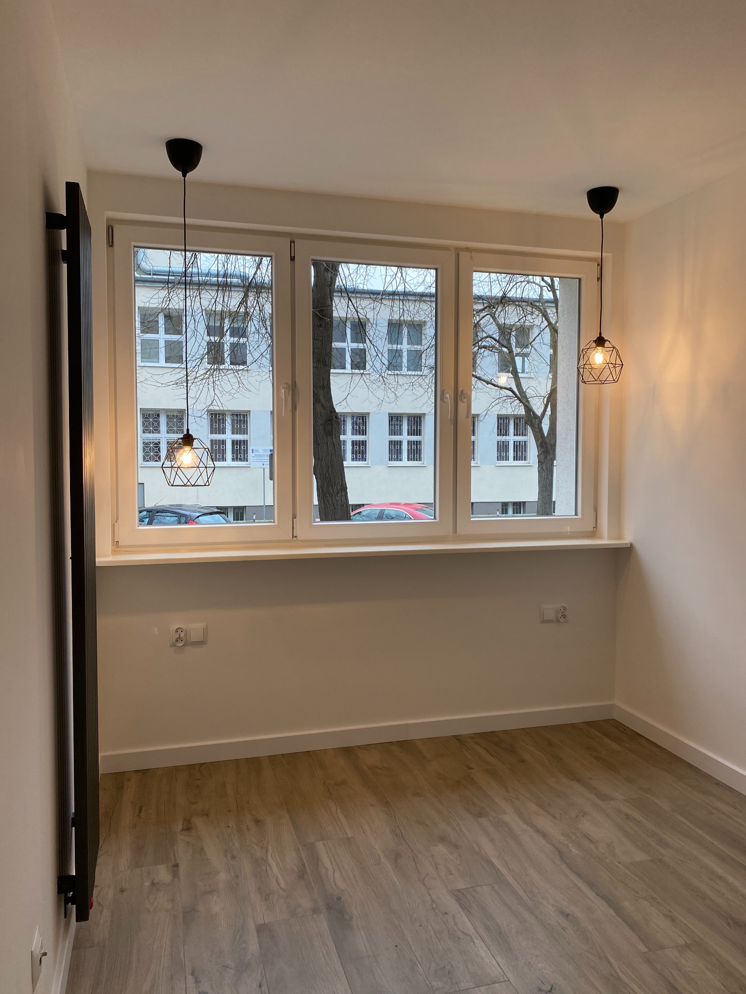 Jasne wnętrze po remoncie: parkiet, okno z widokiem na budynek, nowoczesny grzejnik i dwie lampy wiszące z żarówkami Edisona. Minimalistyczny design.