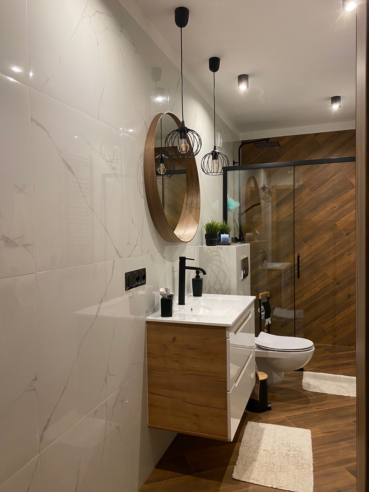 Nowoczesna łazienka z drewnianymi akcentami: szafka pod umywalką, okrągłe lustro, kabina prysznicowa z deseniem drewna i białe płytki imitujące marmur.