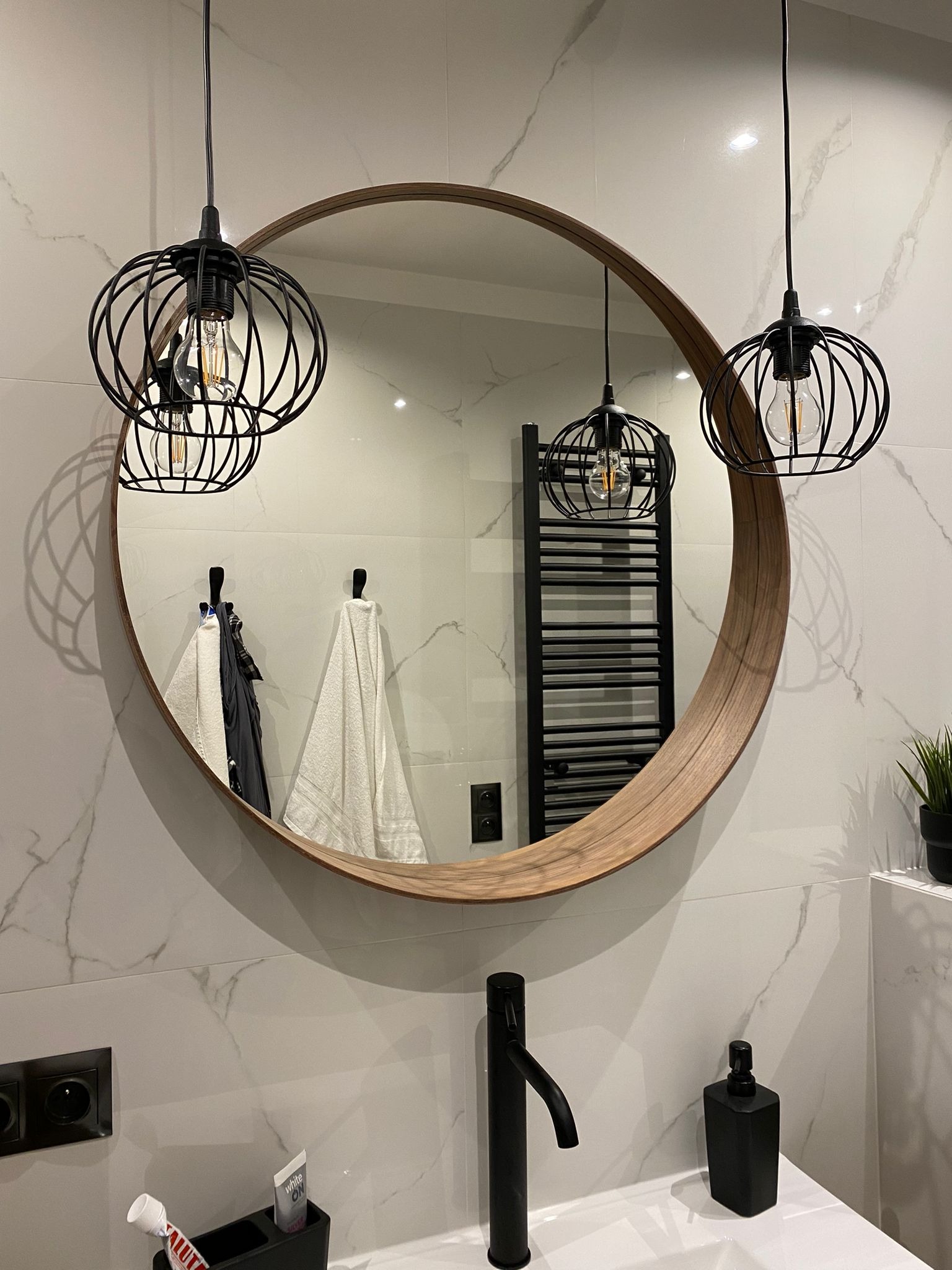 Nowoczesna łazienka z okrągłym lustrem w drewnianej ramie, czarną armaturą i wiszącymi lampami w stylu industrialnym na tle płytek imitujących marmur.