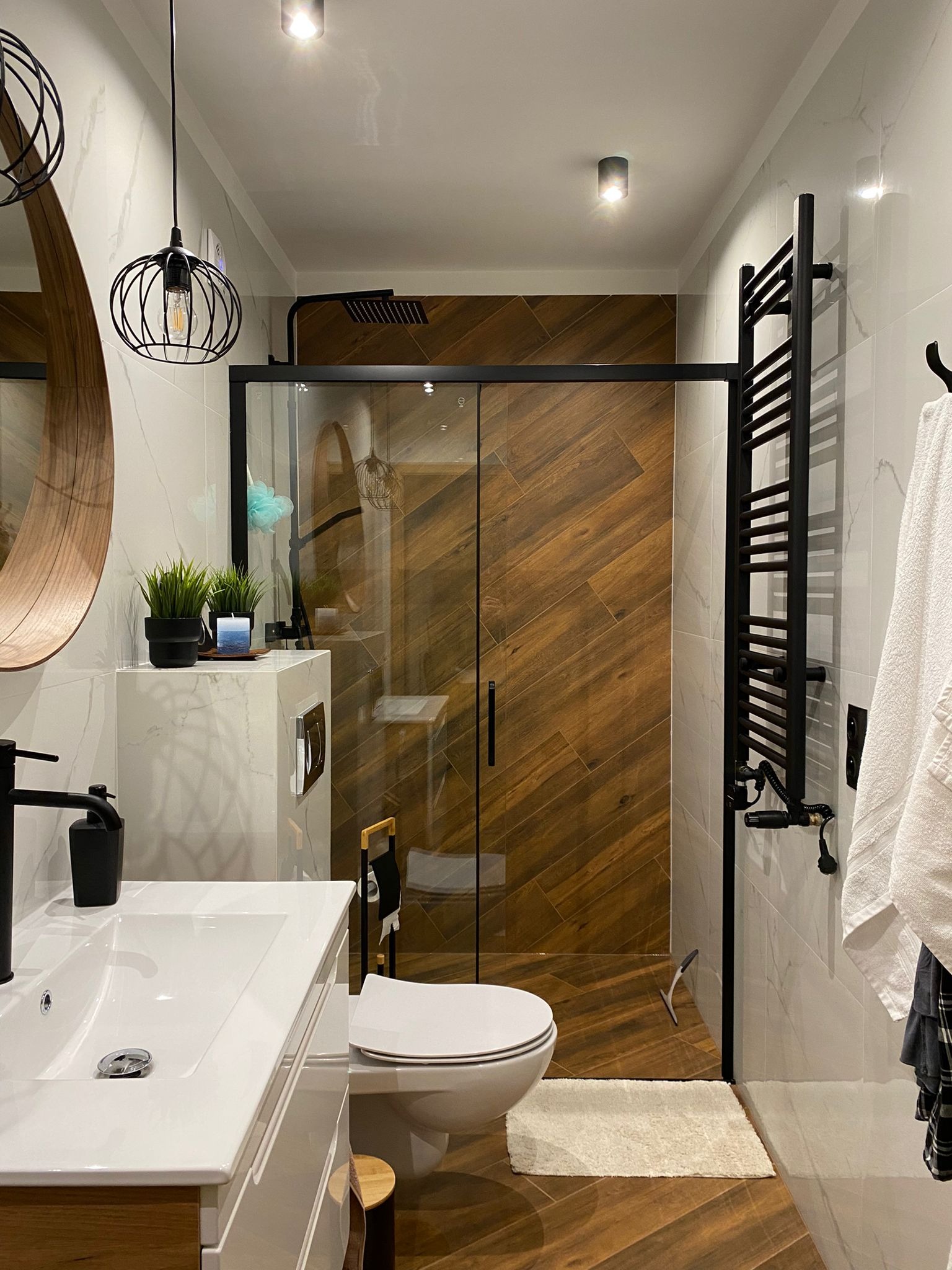 Nowoczesna łazienka w Gdyni z kabiną prysznicową typu walk-in, drewnopodobnymi płytkami ułożonymi w jodełkę i czarnymi akcentami, jak grzejnik i armatura.