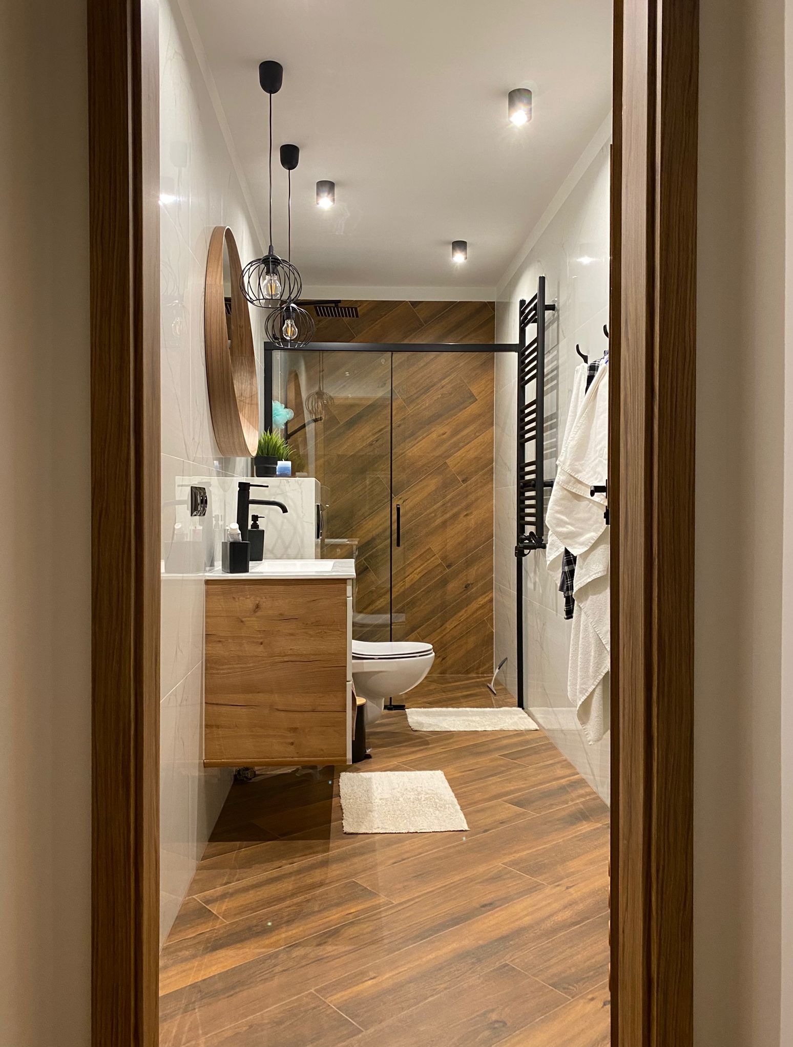 Nowoczesna łazienka z drewnianymi akcentami: płytki imitujące drewno, okrągłe lustro i czarne detale. Kabina prysznicowa ze szklaną ścianą.