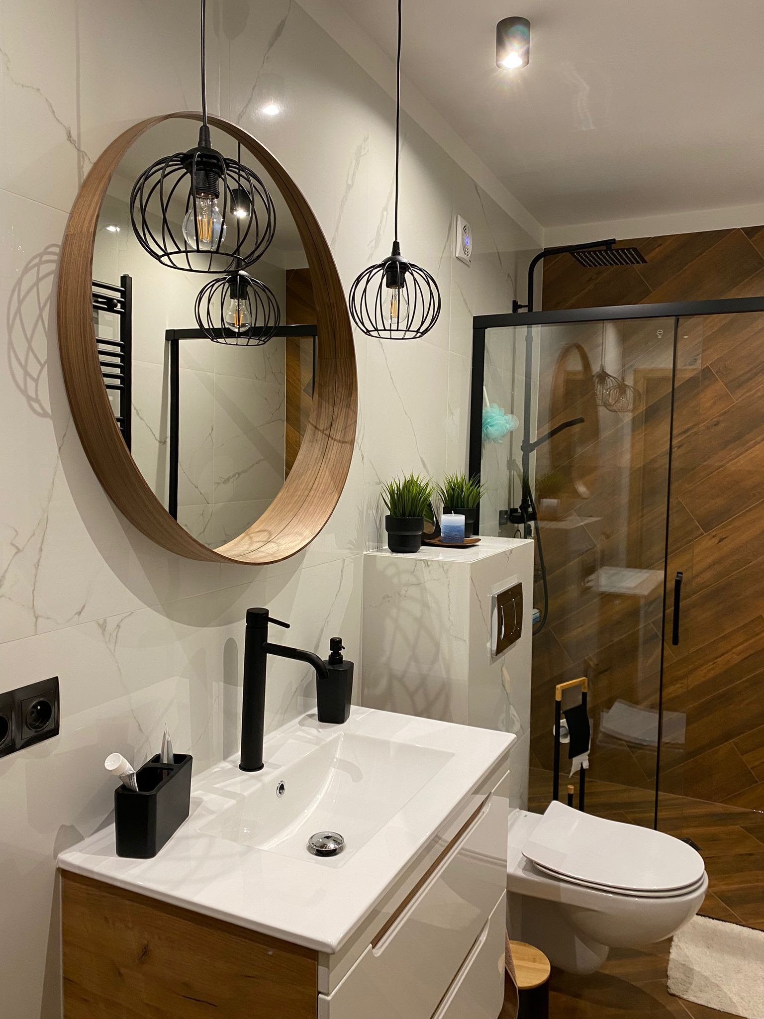 Nowoczesna łazienka z kabiną prysznicową typu walk-in, okrągłym lustrem, wiszącymi lampami w stylu industrialnym i płytkami imitującymi marmur.