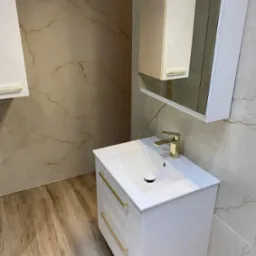 Nowoczesna łazienka z białą szafką pod umywalką, złotą armaturą i lustrem. Płytki imitujące marmur na ścianach i drewnopodobne na podłodze.