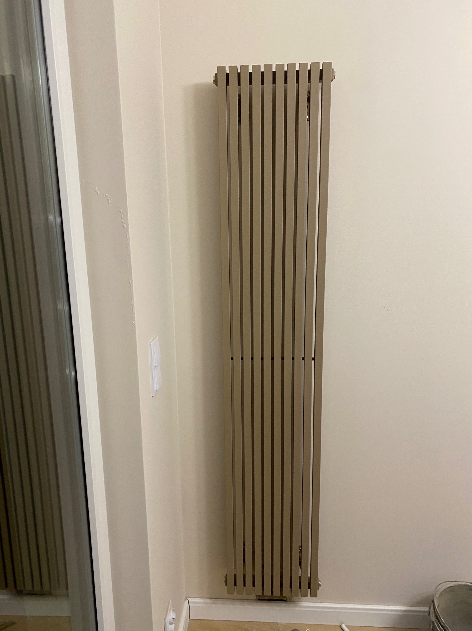 Beżowy, pionowy grzejnik dekoracyjny z pionowymi szczeblami, zamontowany na kremowej ścianie w rogu pomieszczenia, widoczny fragment listwy przypodłogowej i puszka farby.