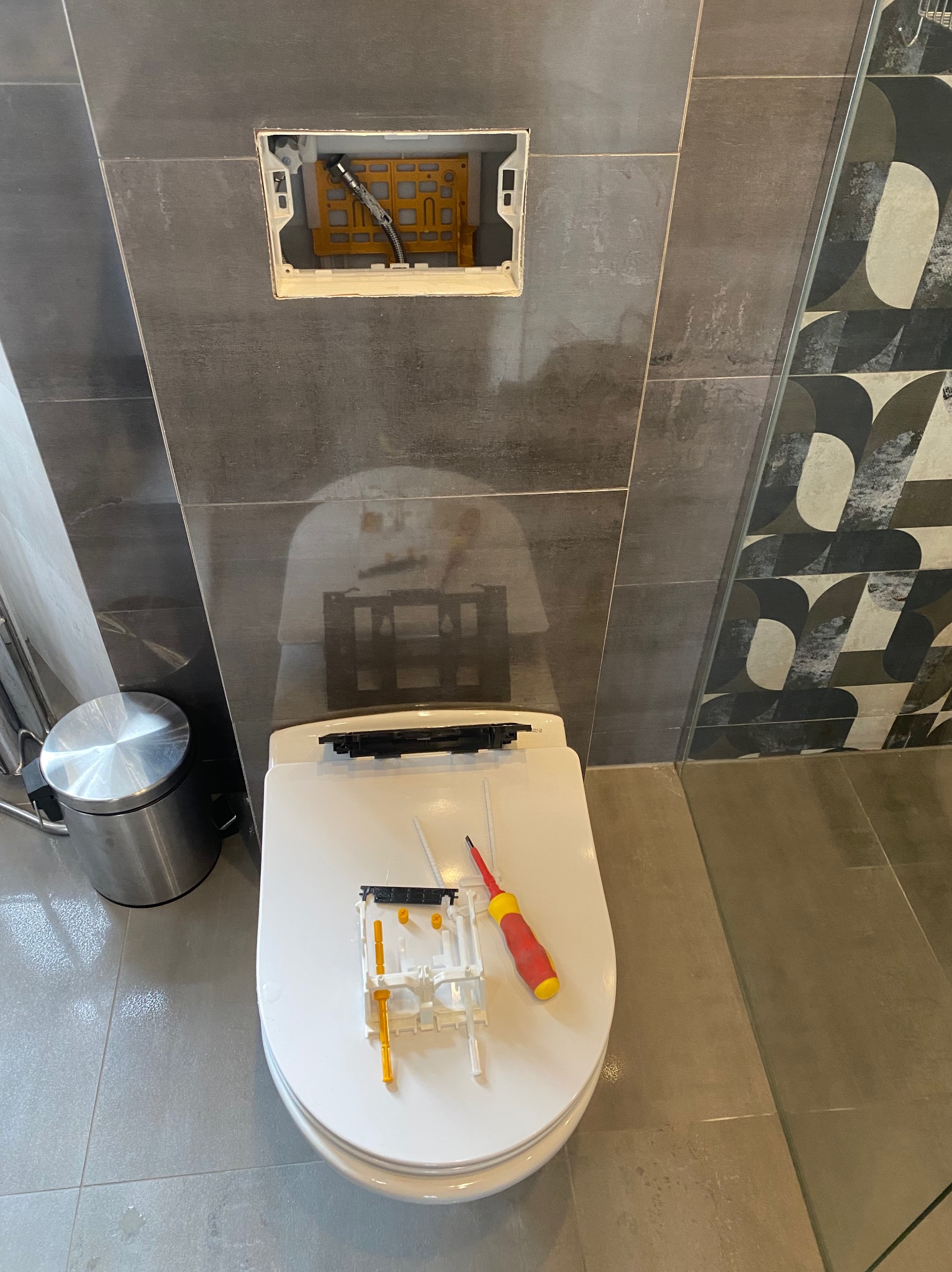 Serwis toalety podtynkowej: widoczne elementy mechanizmu spłukującego na sedesie, narzędzie obok. Tło to szare płytki i wzorzysta dekoracja ścienna.