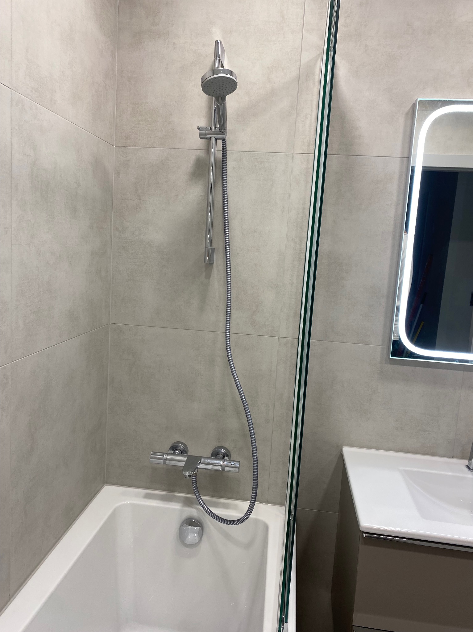 Nowoczesna łazienka z wanną, szklanym parawanem i podświetlanym lustrem. Szare płytki imitujące beton. Chromowana bateria i deszczownica.