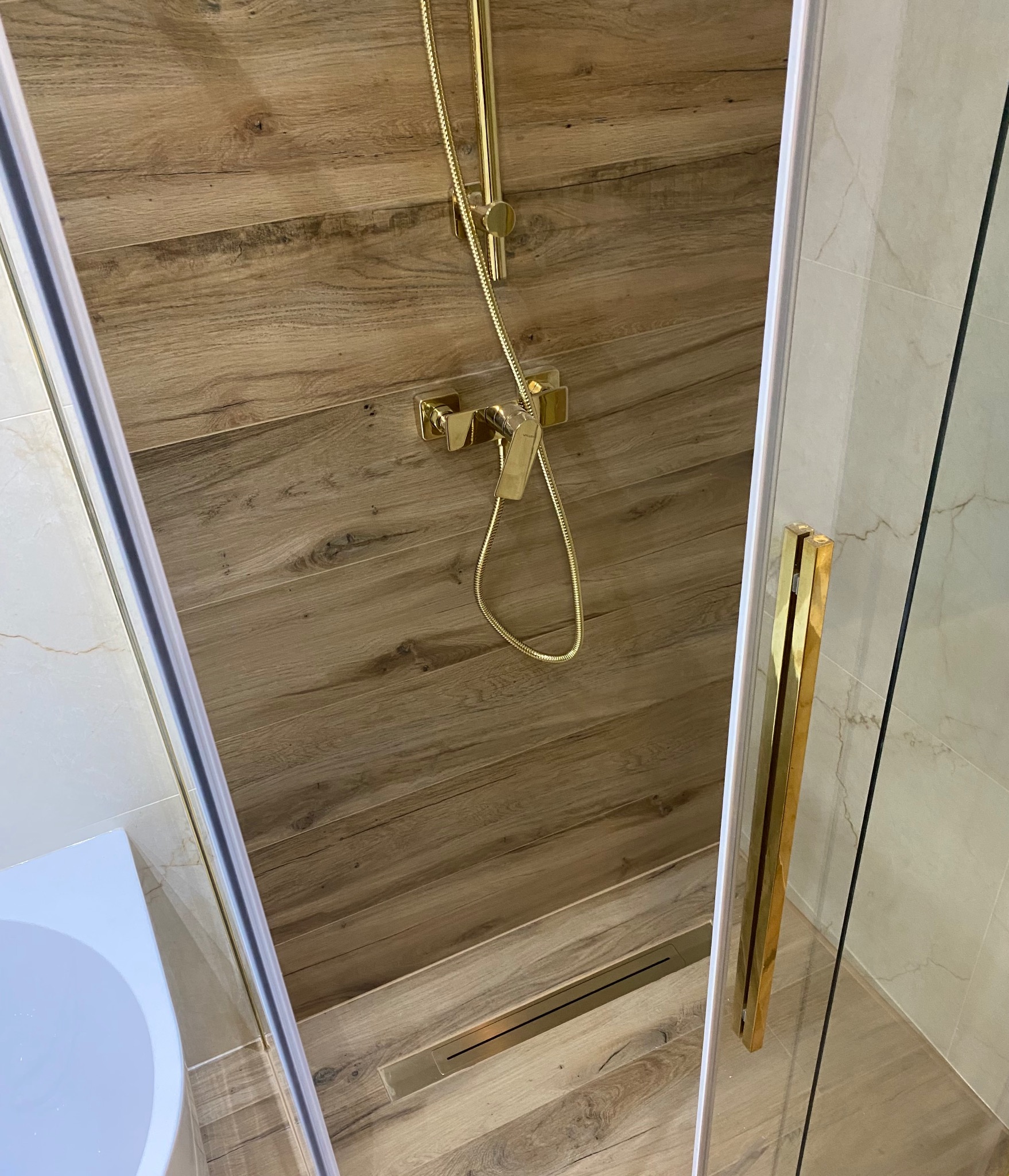 Wykończona łazienka z kabiną prysznicową, złota armatura, drewnopodobne płytki na ścianie i podłodze, odpływ liniowy. Nowoczesny design.