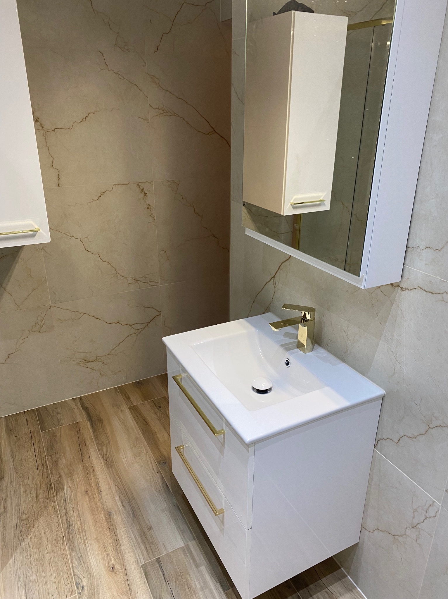 Nowoczesna łazienka z białą szafką pod umywalką, złotą armaturą i lustrem. Płytki imitujące marmur na ścianach i drewnopodobne na podłodze.