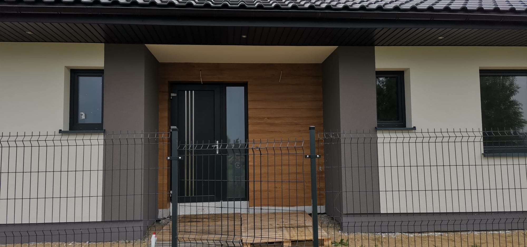 Nowoczesny dom jednorodzinny z elewacją w odcieniach szarości i beżu, z drewnianym akcentem nad wejściem i czarną stolarką okienną. Widok zza ogrodzenia.