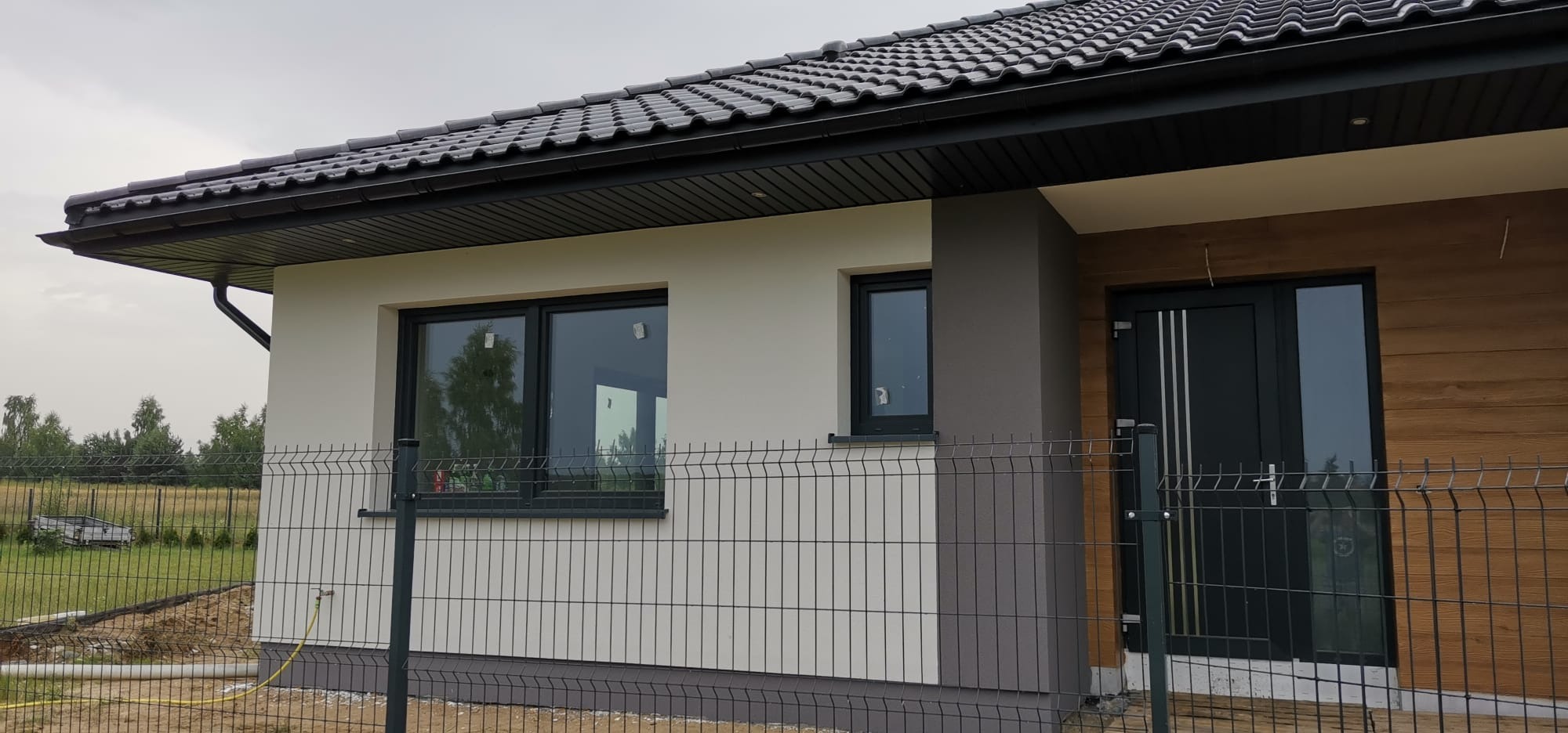 Dom jednorodzinny z elewacją w odcieniach bieli i szarości, widoczny zza ogrodzenia. Nowoczesne okna i drzwi w kolorze antracytowym. Dach pokryty ciemną dachówką.