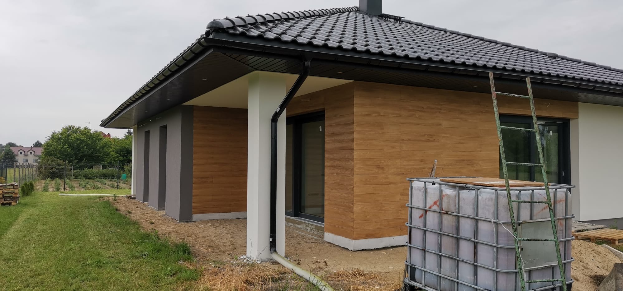 Wykończona elewacja domu jednorodzinnego z drewnopodobną okładziną, widoczne okno i drabina oparta o ścianę. Część elewacji pomalowana na szaro.