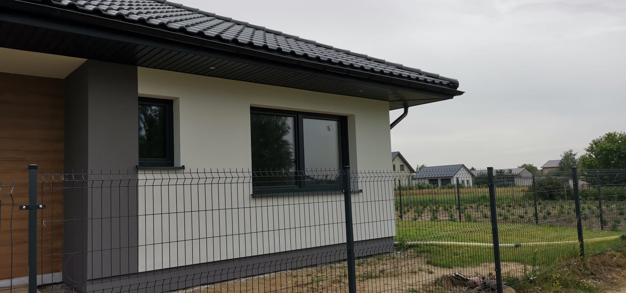 Fragment domu jednorodzinnego z elewacją w odcieniach bieli i szarości, widoczny przez metalowe ogrodzenie. Dach pokryty czarną dachówką.