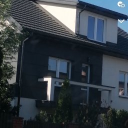Daniel Groszyk - Dwukondygnacyjny dom z ciemnoszarą elewacją części parteru i białymi ścianami na piętrze, ciemna dachówka, komin i antena na dachu, widok zza ogrodzenia.