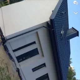 Nowoczesny dom z ciemną dachówką i jasną elewacją, widok z dołu, z widocznymi oknami i elementami wykończeniowymi.