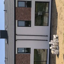 Nowoczesny dom z grafitowym dachem, jasną elewacją z fragmentami cegły klinkierowej wokół okien, rynnami i stertą białych bloczków na ziemi.