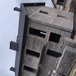 Daniel Groszyk - Surowy budynek w trakcie wznoszenia z szarej cegły, z widoczną konstrukcją dachu pokrytą ciemną dachówką, palety z cegłami obok, pochmurne niebo w tle.