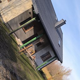 Daniel Groszyk - Nowoczesny dom z ciemnym dachem z dachówki, wyposażonym w komin i okno połaciowe, na tle błękitnego nieba. Widoczna jasna elewacja i ciemne rynny.