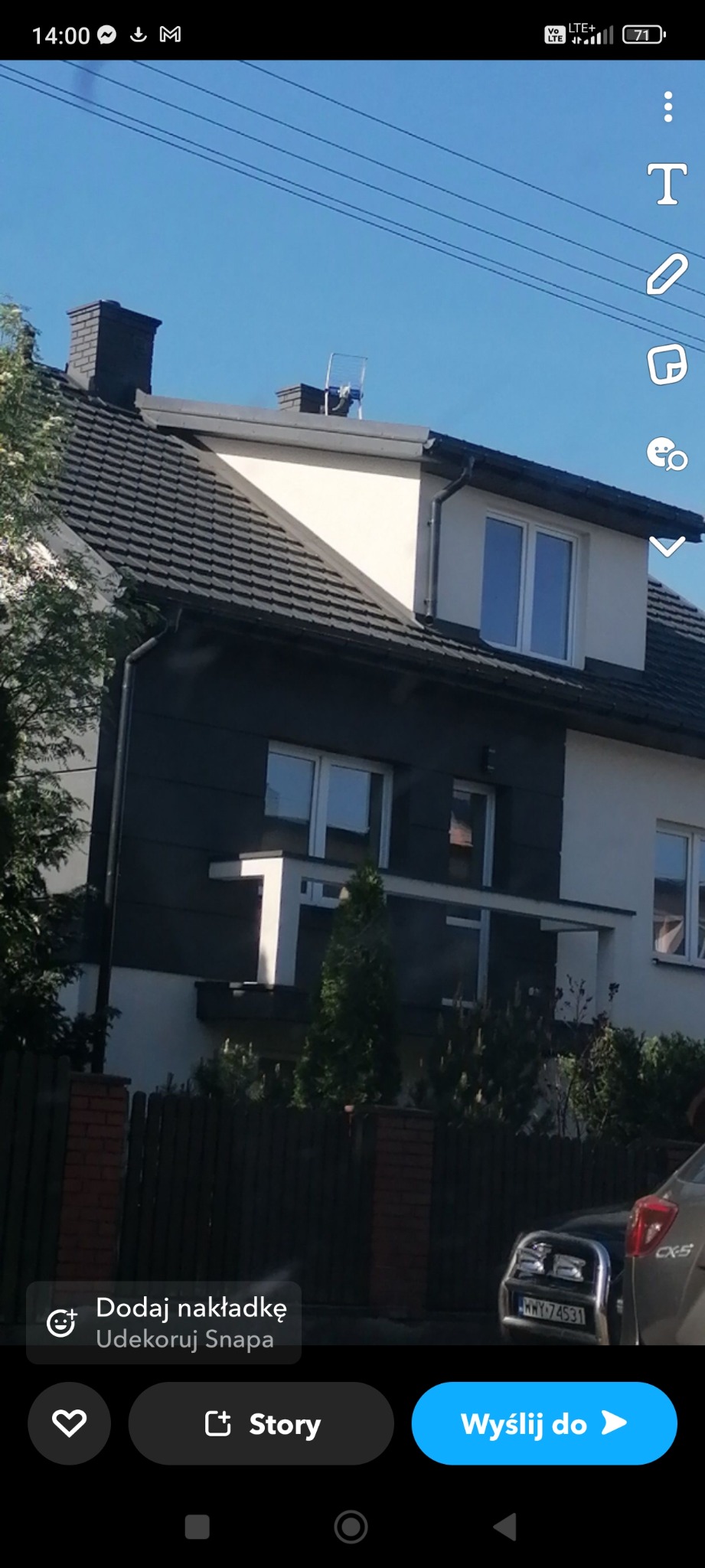 Dwukondygnacyjny dom z ciemnoszarą elewacją części parteru i białymi ścianami na piętrze, ciemna dachówka, komin i antena na dachu, widok zza ogrodzenia.