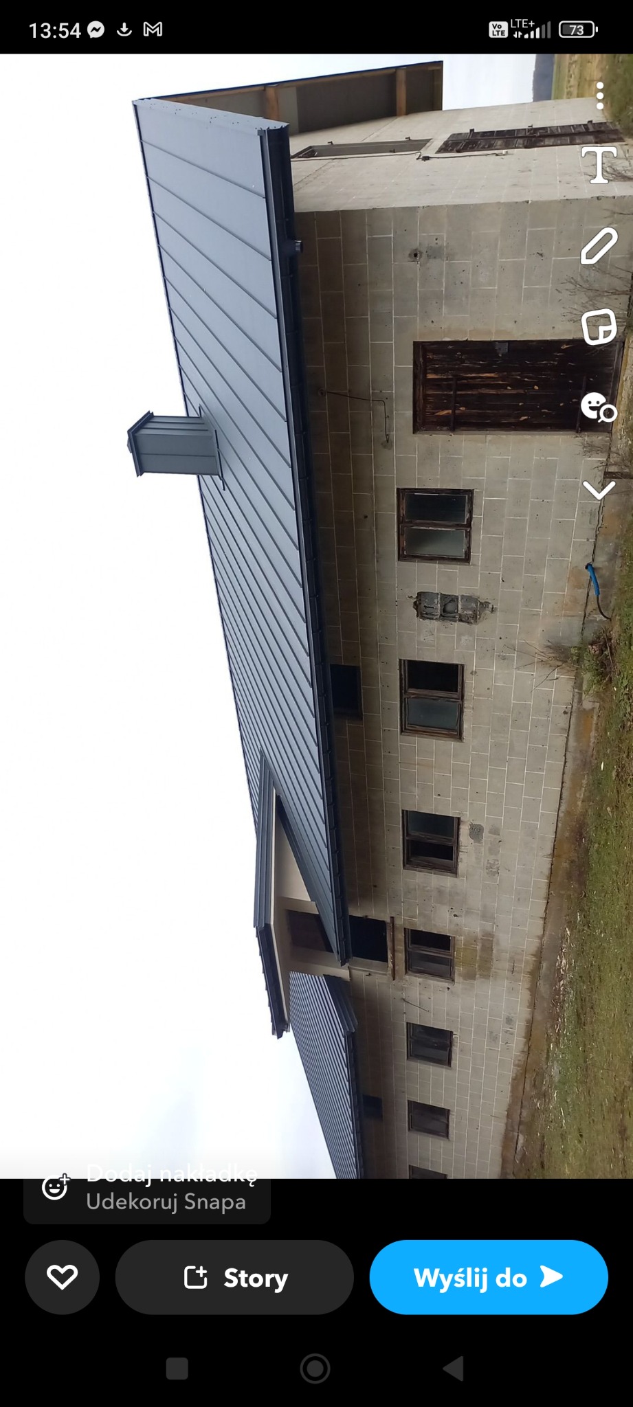 Szara blacha na dachu budynku z szarego pustaka, widoczny fragment elewacji i małe okna z drewnianymi okiennicami.