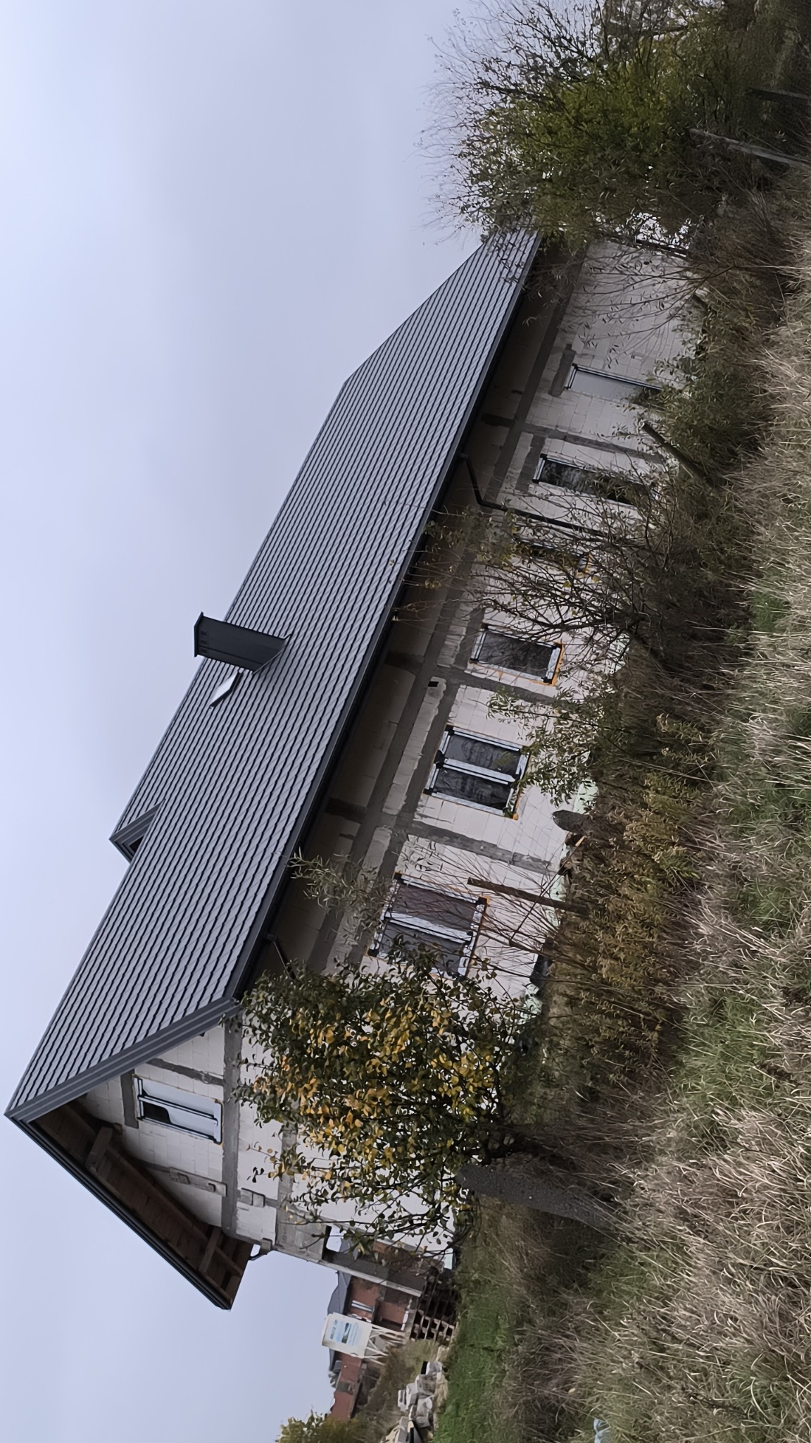 Dom w trakcie budowy z nowym, ciemnoszarym dachem z blachy trapezowej i widoczną konstrukcją ścian z pustaków, otoczony wysoką trawą i krzewami w jesiennej scenerii.