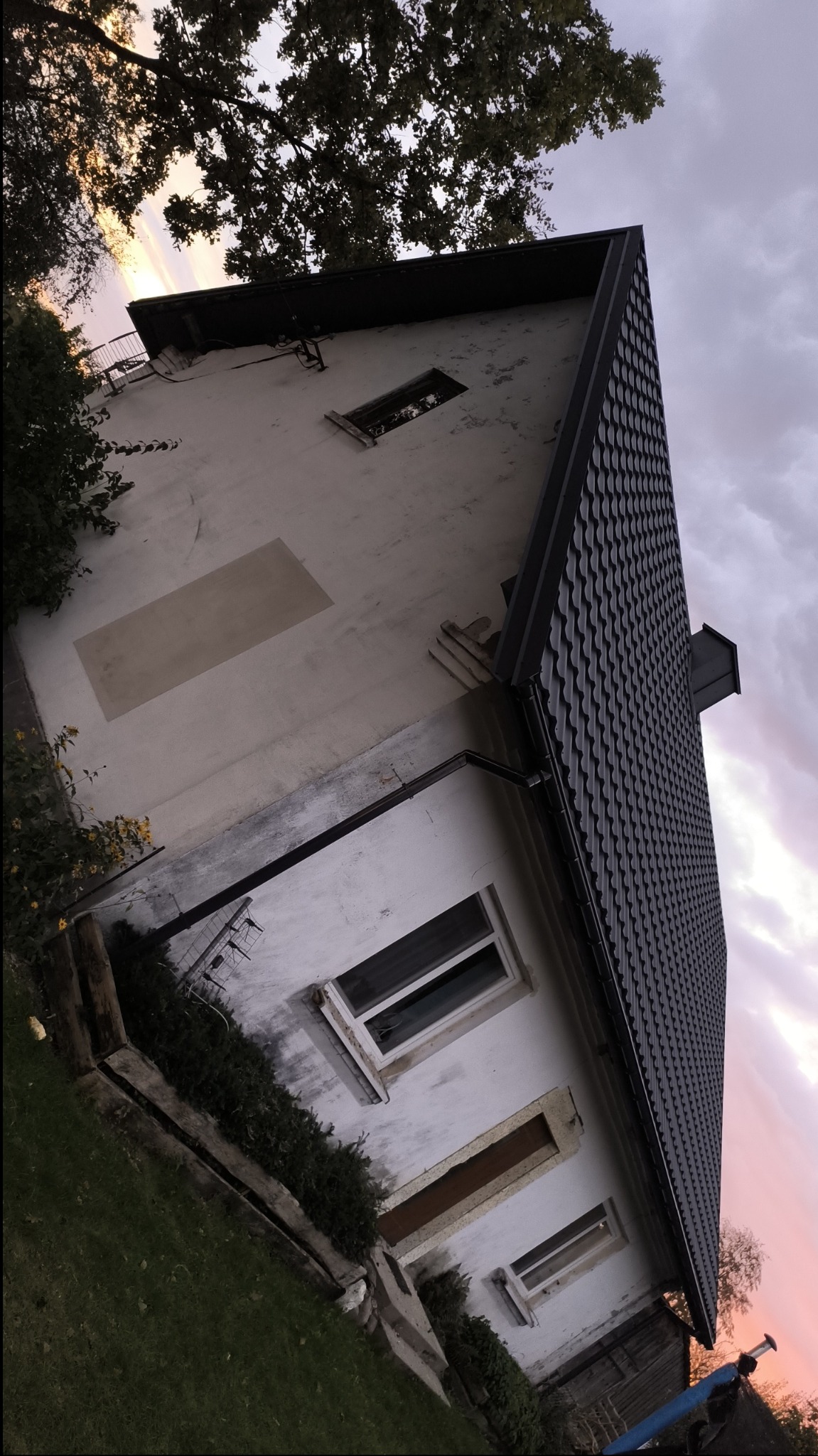 Dom jednorodzinny z nowym, czarnym dachem z blachodachówki, widok z dołu na tle zachodzącego słońca i pochmurnego nieba. Na elewacji widoczne ślady po naprawach.