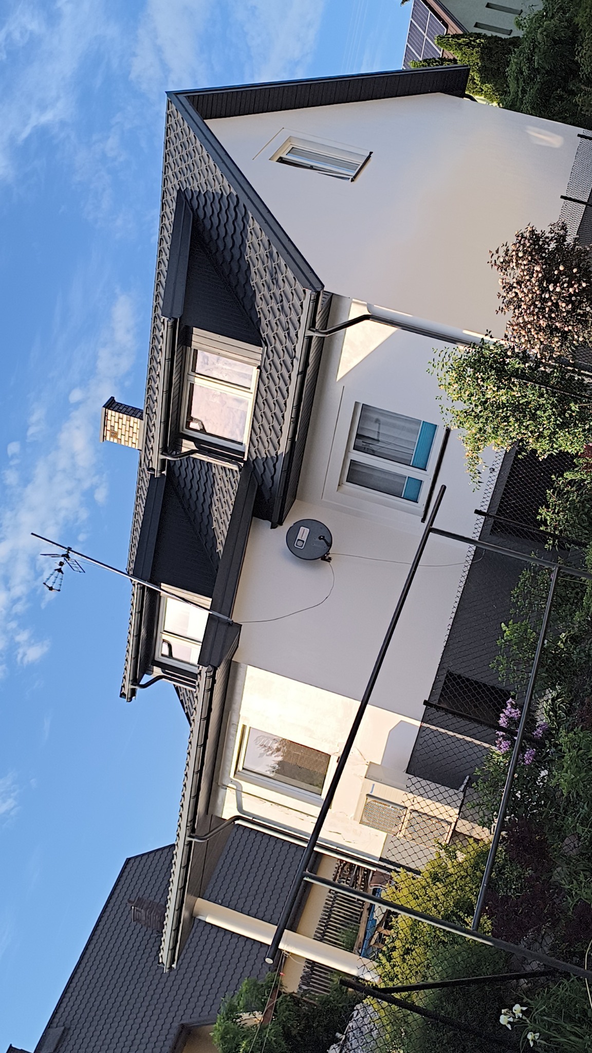 Elewacja domu z ciemną dachówką, widoczne okna z białymi ramami, antena satelitarna i fragment ogrodu z siatką ogrodzeniową.