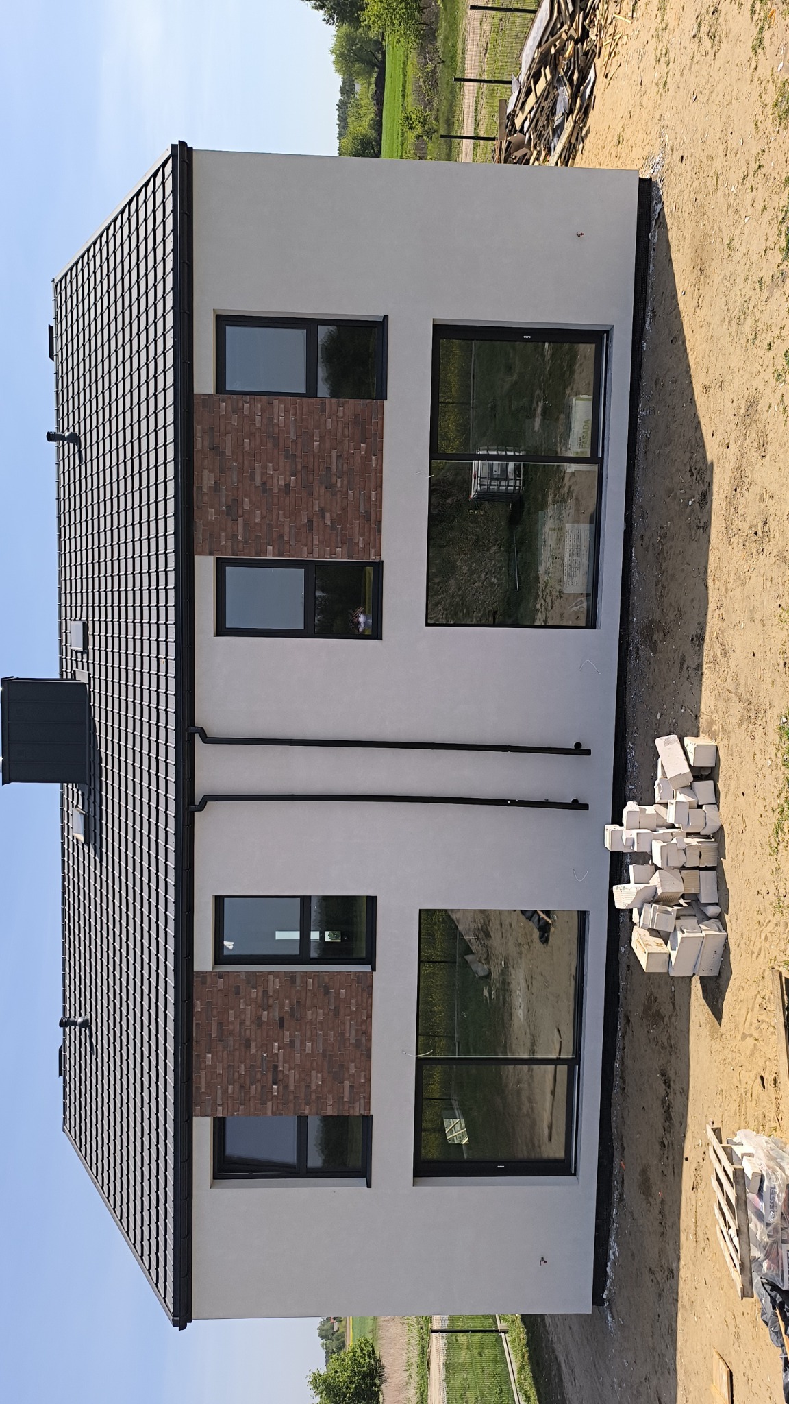 Nowoczesny dom z grafitowym dachem, jasną elewacją z fragmentami cegły klinkierowej wokół okien, rynnami i stertą białych bloczków na ziemi.