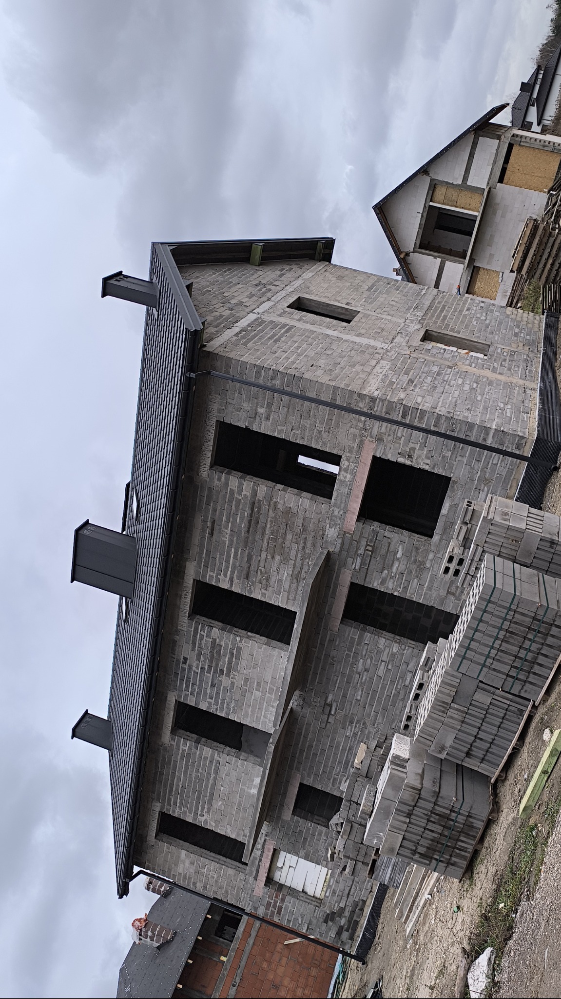 Surowy budynek w trakcie wznoszenia z szarej cegły, z widoczną konstrukcją dachu pokrytą ciemną dachówką, palety z cegłami obok, pochmurne niebo w tle.