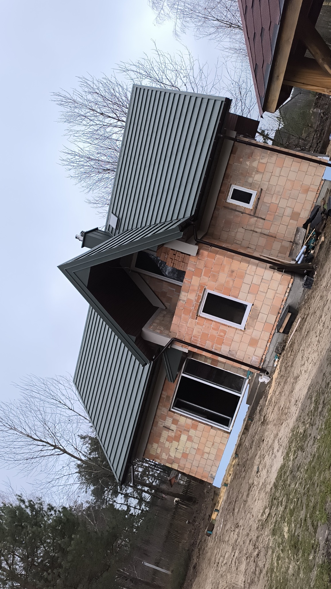 Dom w trakcie budowy w Długosiodle z nowym, zielonym dachem z blachy trapezowej i czerwoną elewacją z cegły klinkierowej, okna bez szyb, widoczne rusztowania i materiały budowlane.