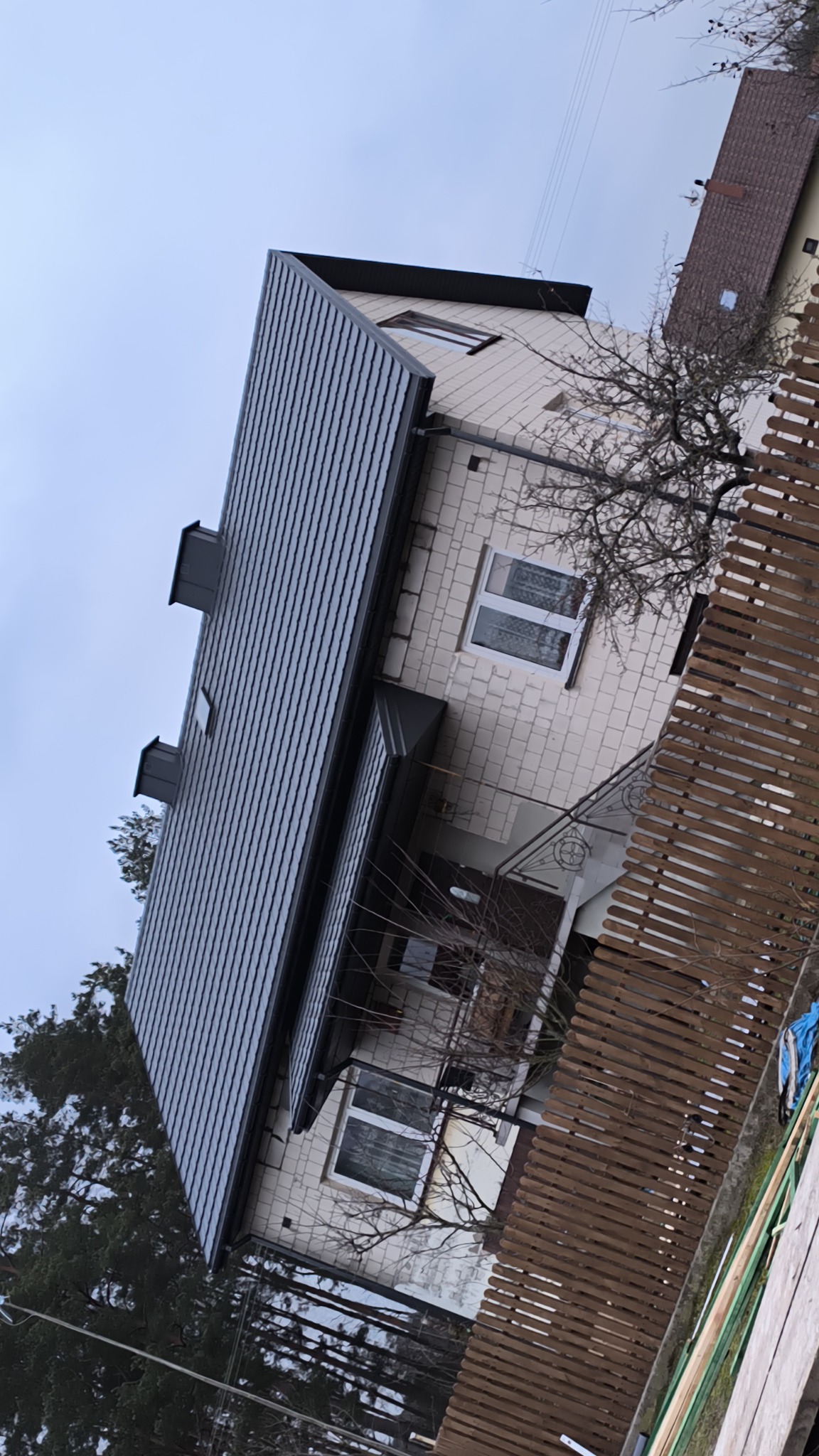 Dom jednorodzinny z nowym, ciemnoszarym dachem z blachy trapezowej, widoczne kominy i okno dachowe, elewacja z jasnej cegły, ujęcie z dołu zza drewnianego płotu.